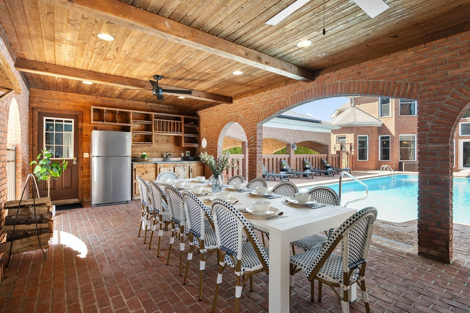 Outdoor covered patio with a brick floor, wooden ceiling, and brick walls. There is a dining table set with white bowls and plates, and a floral centerpiece. The patio overlooks a backyard with lounge chairs, a swimming pool, and a brick house in the