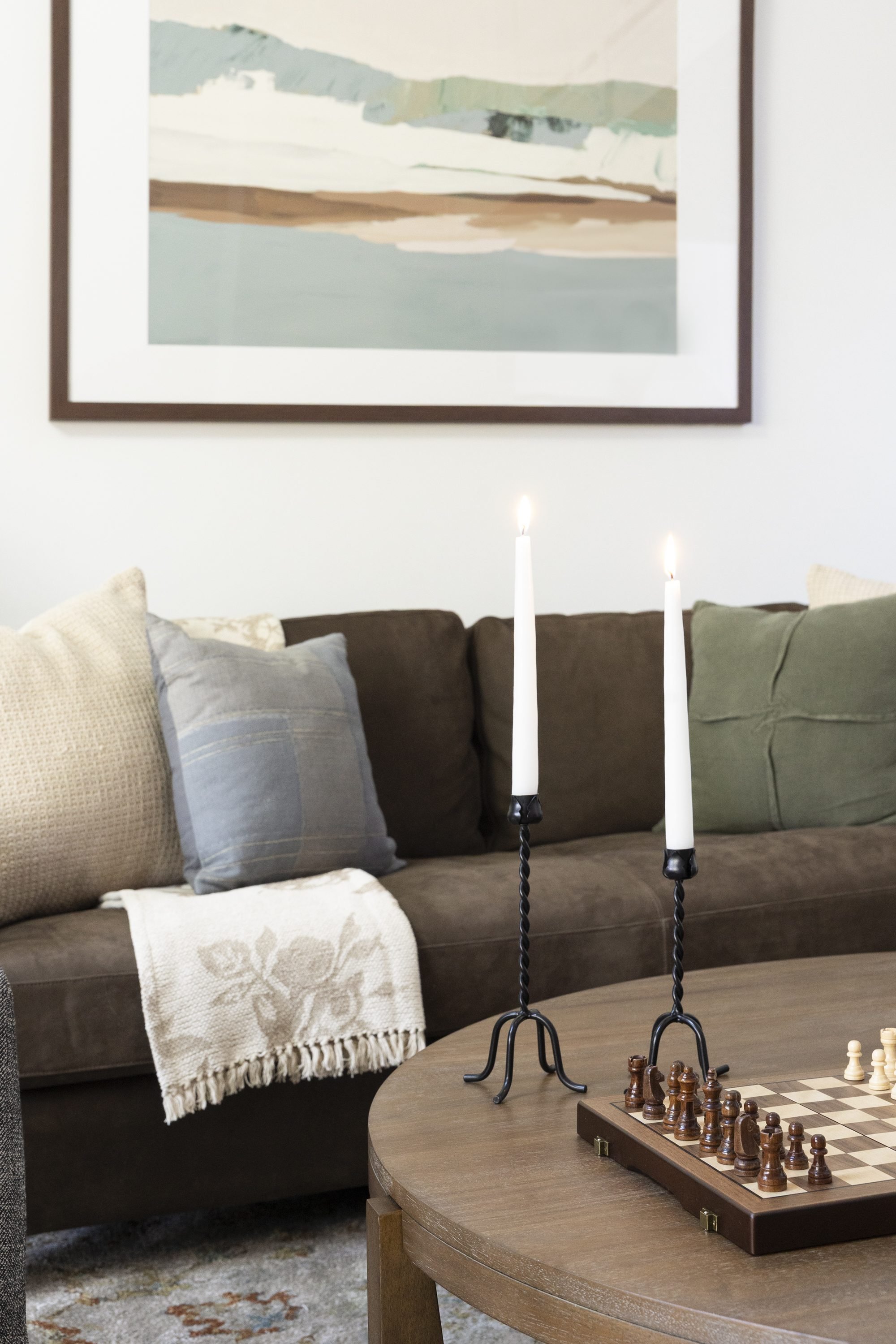 Living room with a dark brown sofa, beige and gray pillows, a white throw blanket, a painting on the wall, a wooden table with two lit candles, and a chessboard.