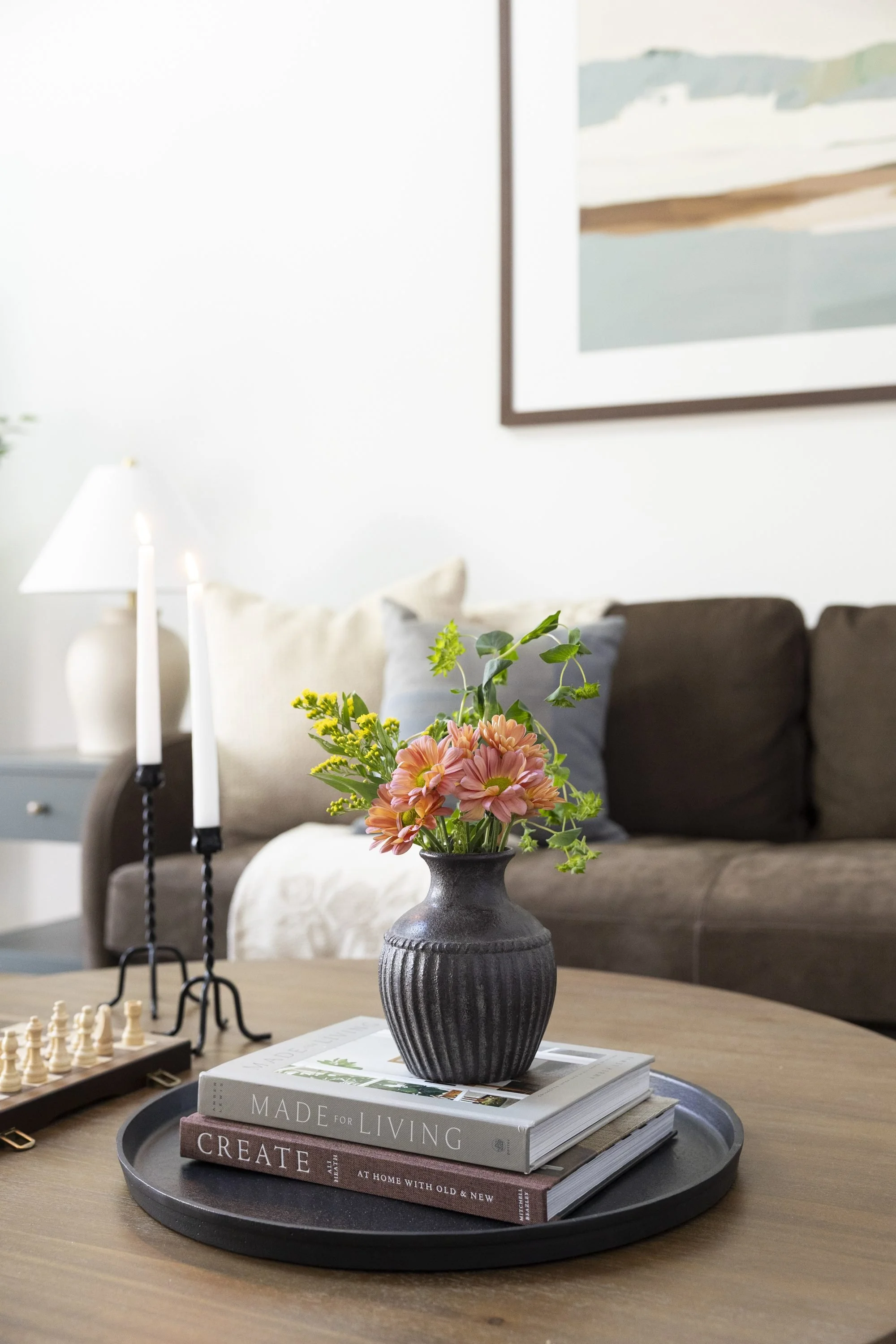 A black ceramic flower vase with pink and yellow flowers and green foliage on top of a stack of books on a tray, on a round wooden table in a cozy living room.