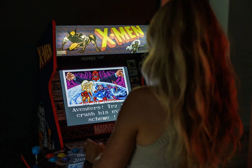 A woman with long hair is playing an X-Men arcade game in a dark room.