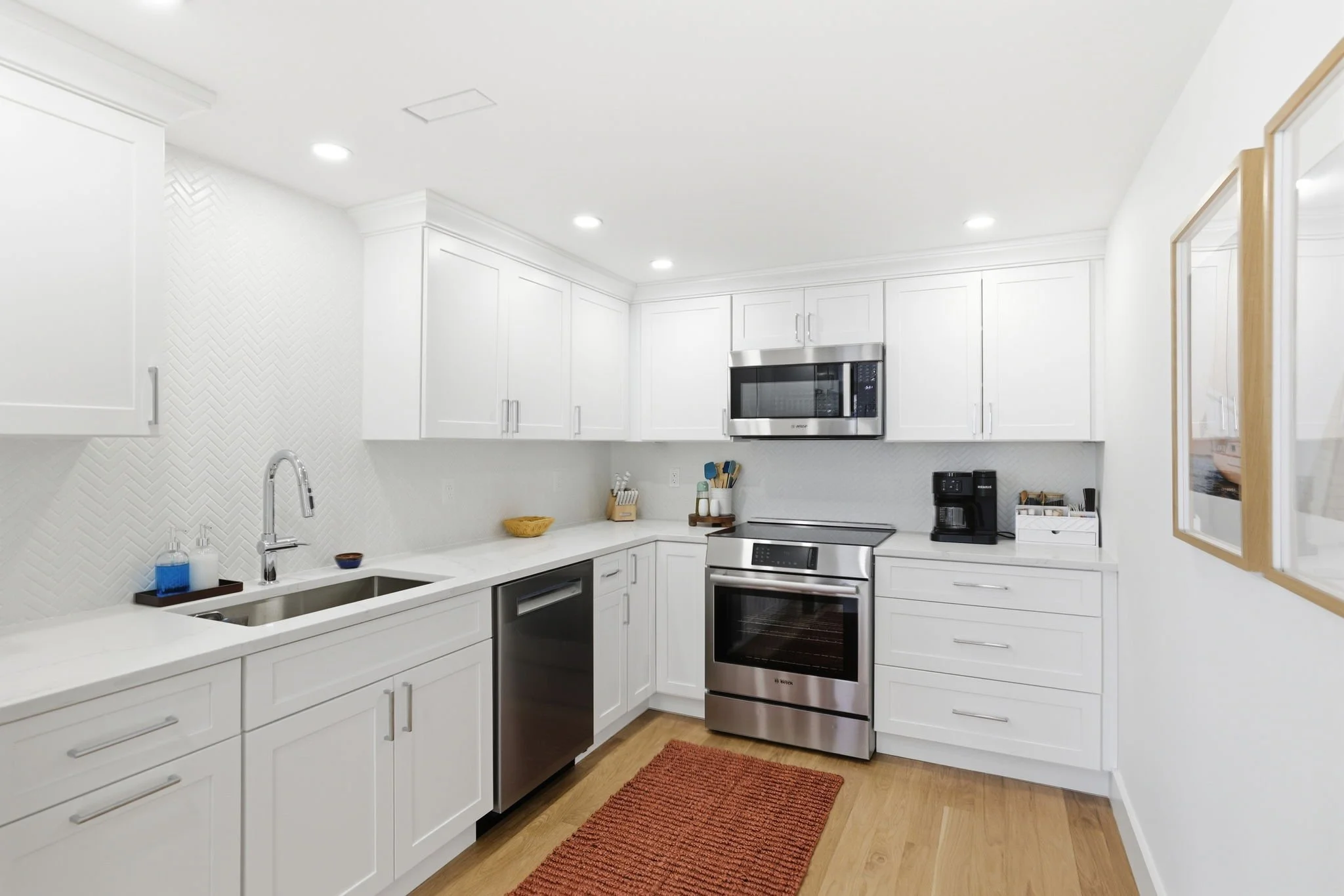 A modern kitchen with white cabinets, a stainless steel oven, microwave, and dishwasher, a white countertop with kitchen utensils, a coffee maker, and a small framed picture on the wall, illuminated by ceiling lights and natural light from a window.