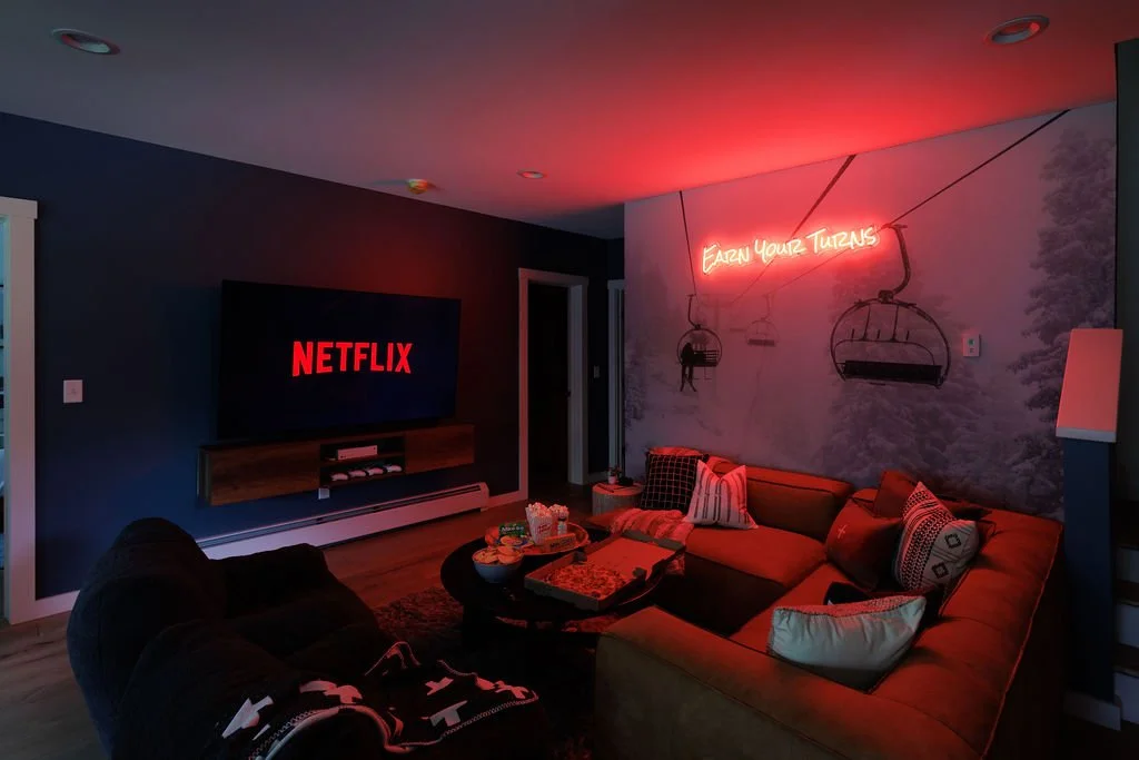 Cozy living room with dark walls, neon sign reading 'Earn Your Turns', a large flat-screen Netflix display, and a sectional sofa with pillows, illuminated by red and soft lighting.