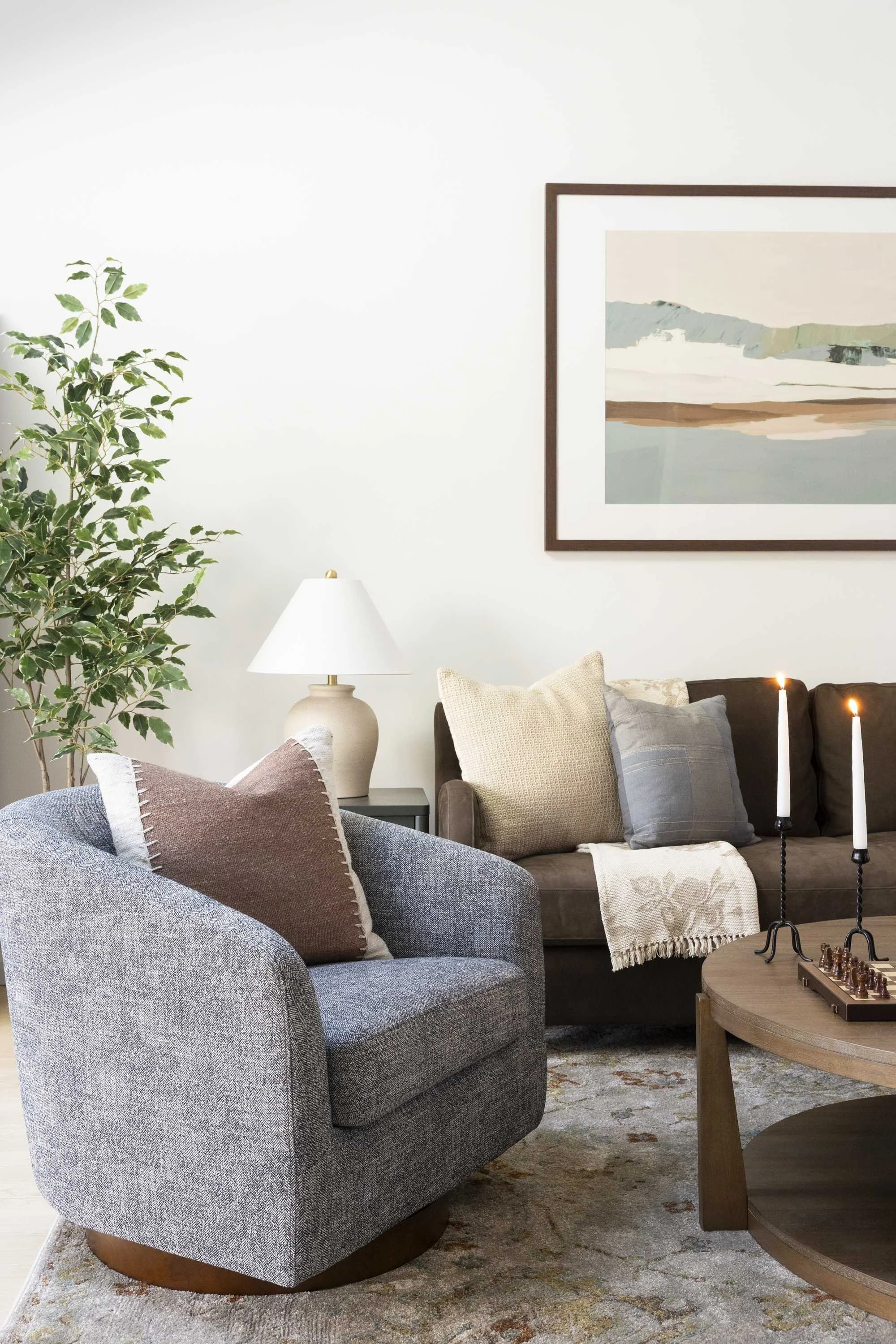Living room with gray patterned armchair, beige loveseat with various cushions, wooden coffee table with candles and chessboard, potted green plant, white lamp, framed landscape painting on white wall, and a patterned area rug.