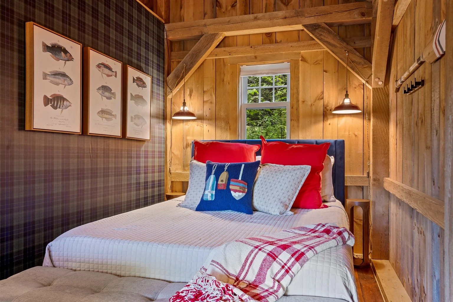 A cozy bedroom with wooden walls and ceiling, a window with greenery outside, a bed with white quilt, red pillows, and decorative pillows with hanging ornaments, flanked by hanging lamps.