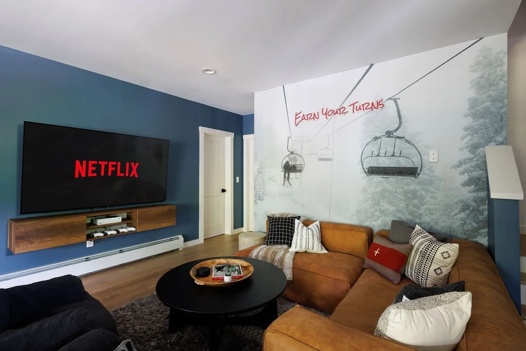 Living room with a large wall mural of gondolas on a snowy mountain, some with people, and red writing saying "Eden your turns." A flat-screen TV displaying "Netflix" is mounted on a blue accent wall. A brown sectional sofa with patterned pillows, a 
