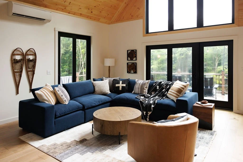 Modern living room with large L-shaped navy sofa, tan armchair, wooden coffee table, and large windows showing outdoors, wood-paneled ceiling, framed wall decor, and an air conditioning unit.