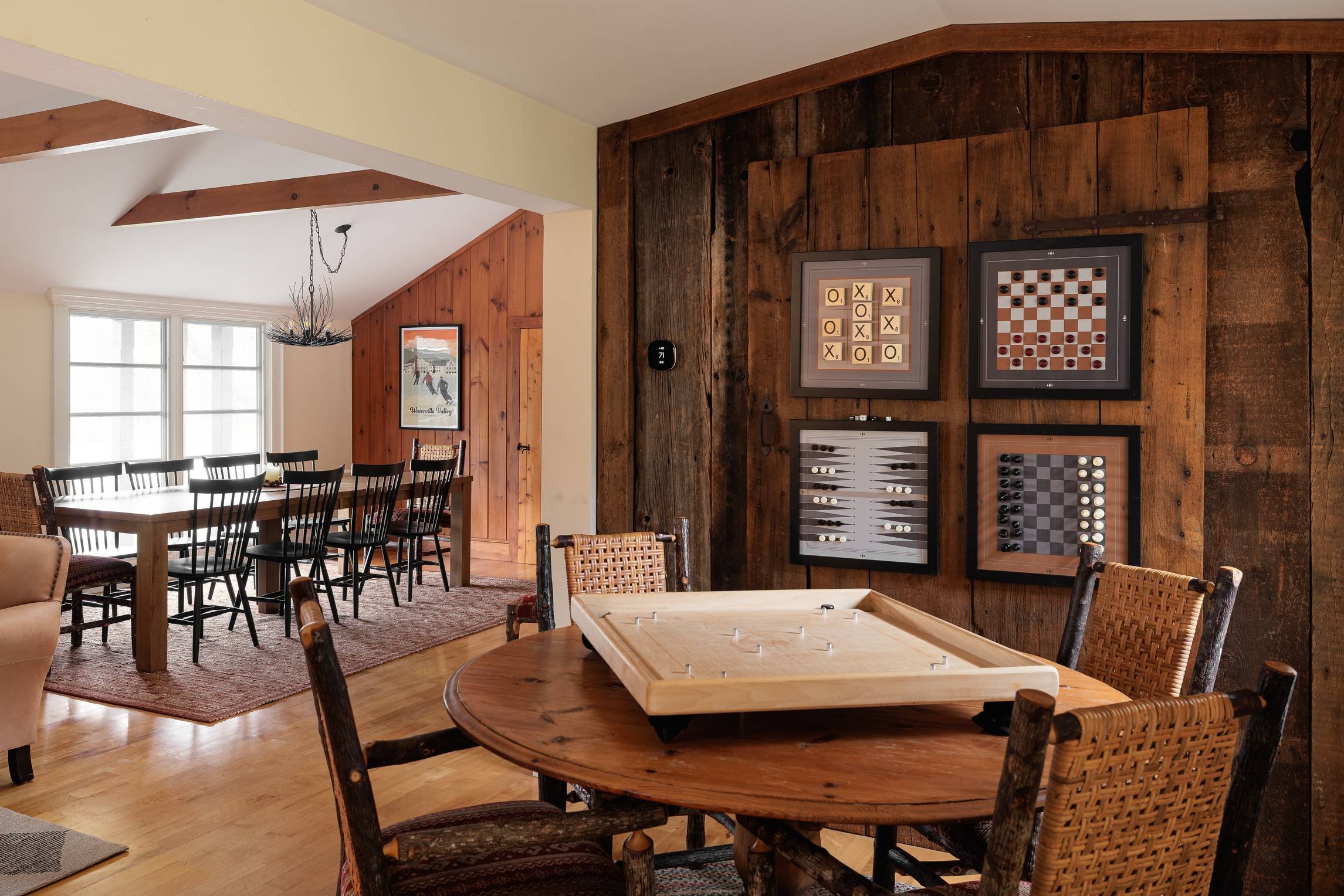 A cozy dining room with wooden furniture, a rectangular table, and multiple chairs. There is a wooden wall with three framed game board pictures and a clock. A window with natural light shines in, and a modern chandelier hangs from the ceiling.