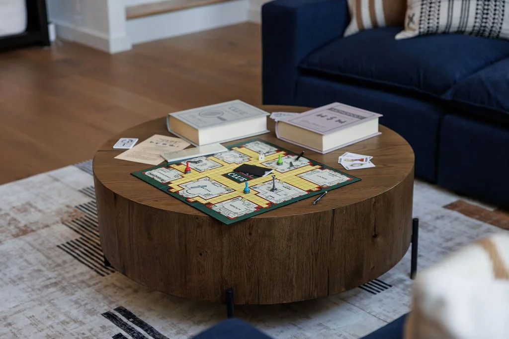 A round wooden coffee table with a Clue board game set up on it, pieces placed on the board, and two large unopened books or manuals nearby. The table is in a living room with a blue sofa and a white rug with black and beige stripes.