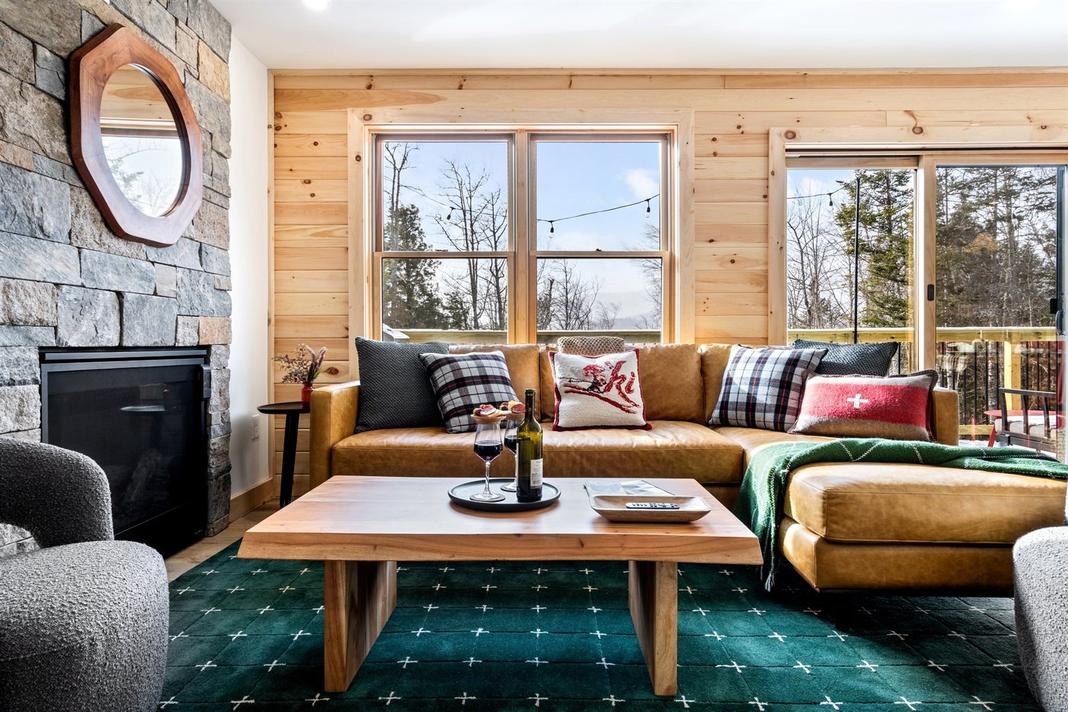 Living room with a tan leather sofa, patterned cushions, a wooden coffee table with wine and snacks, a green throw blanket, a stone fireplace, large windows showing a winter landscape, and a sliding door leading to a balcony.