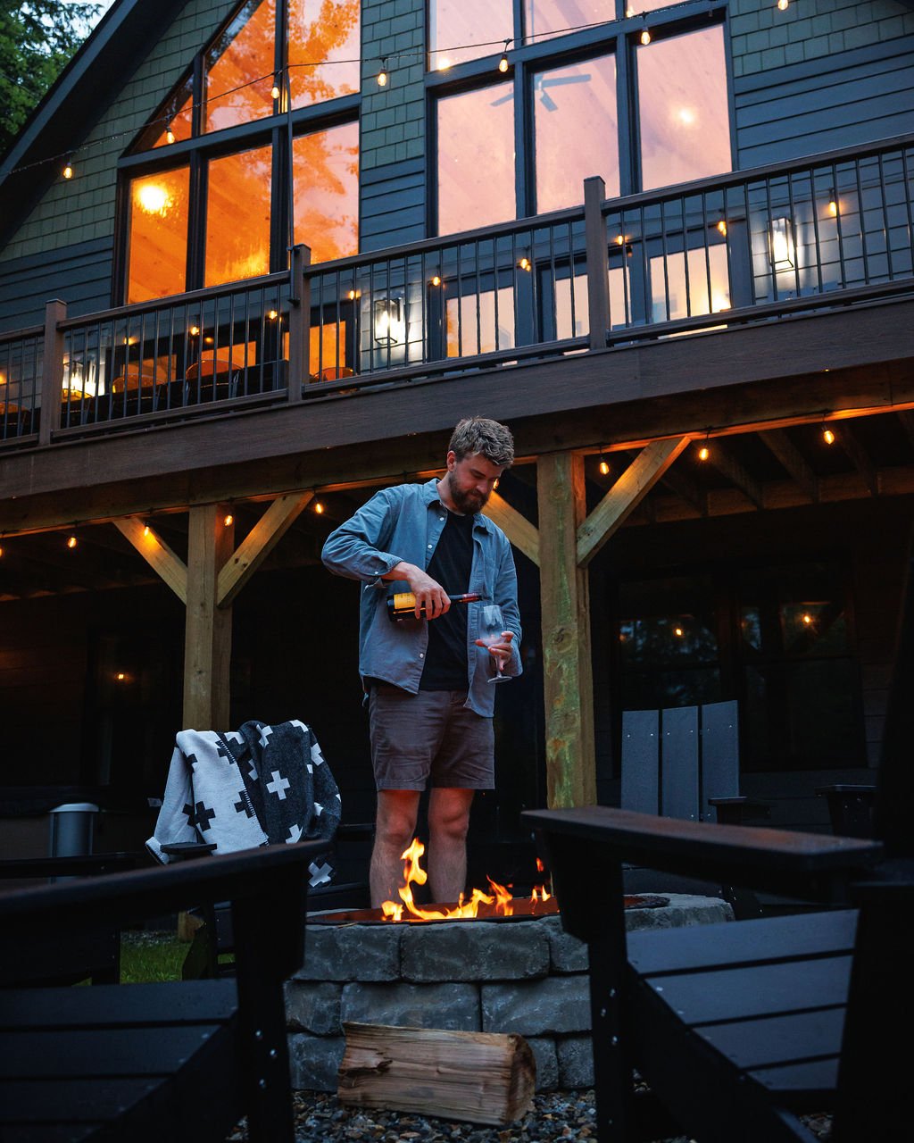 A man pouring wine into a glass in a backyard during sunset, with a fire pit in the foreground and a house with large windows and a balcony in the background.