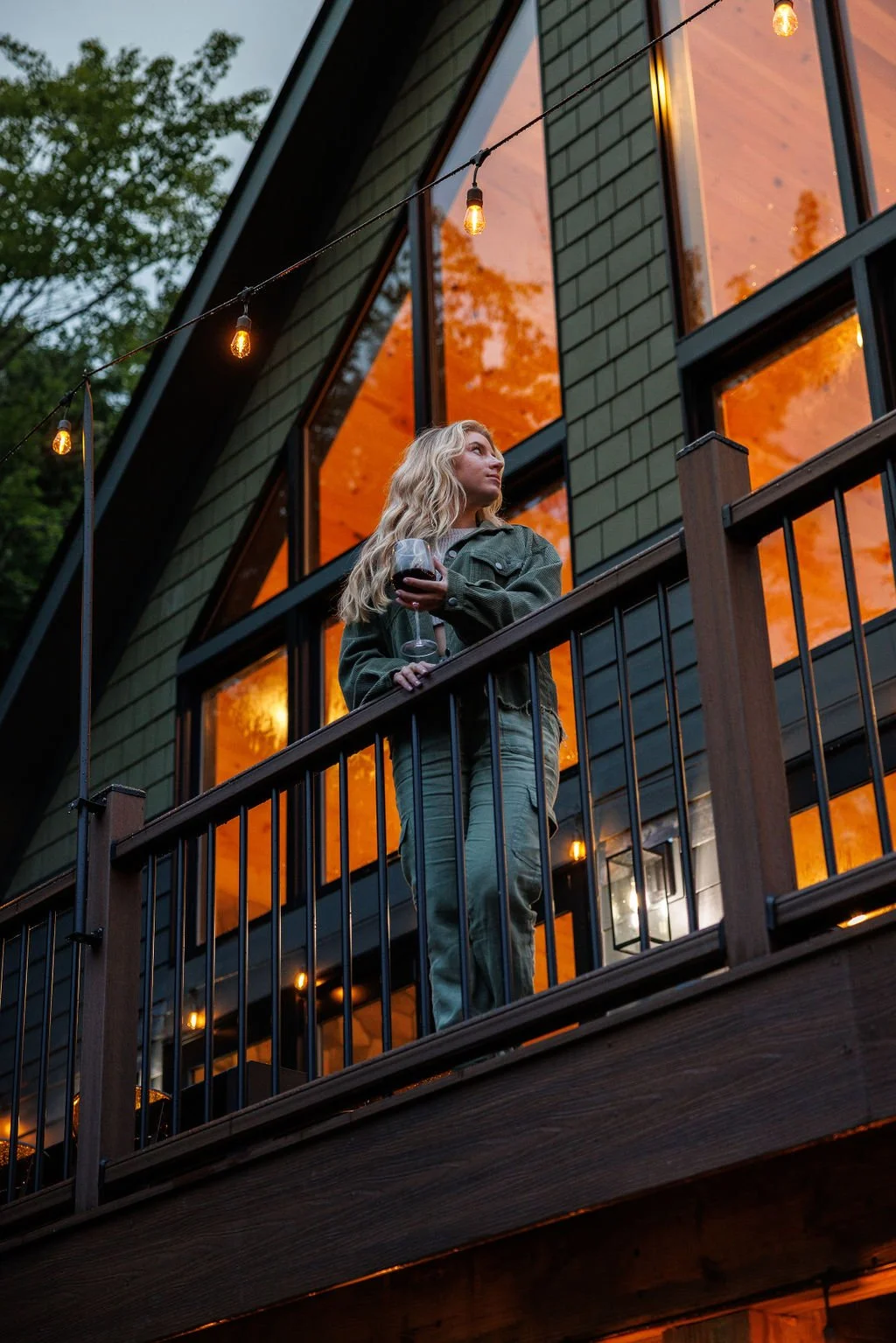 A woman with long blonde hair holding a glass of red wine on a balcony at dusk, with warm outdoor string lights and a modern house with large windows reflecting orange sunset colors.