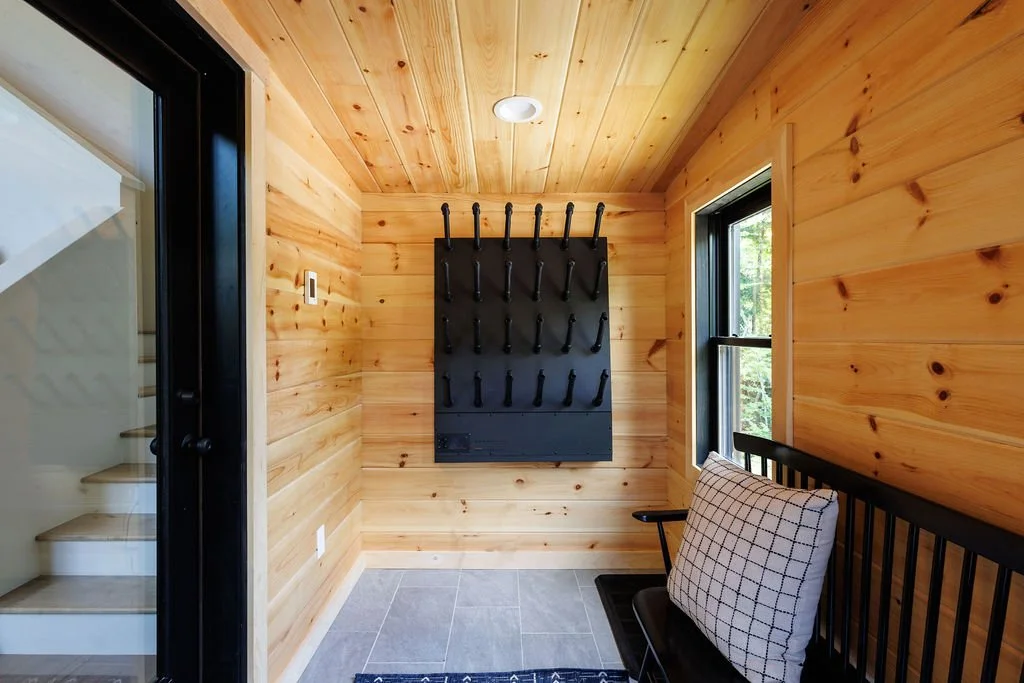 A small room with wood-paneled walls and ceiling, a window showing trees outside, a black wall-mounted rack with hooks, a black chair with a checkered cushion, and a staircase partially visible through a glass door.