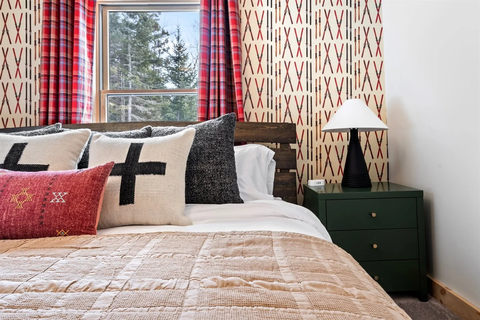 A bedroom with a large window showing trees outside, patterned wallpaper with crossed pairs of skis, red plaid curtains, a bed with multiple decorative pillows, and a dark green nightstand with a lamp and an alarm clock.
