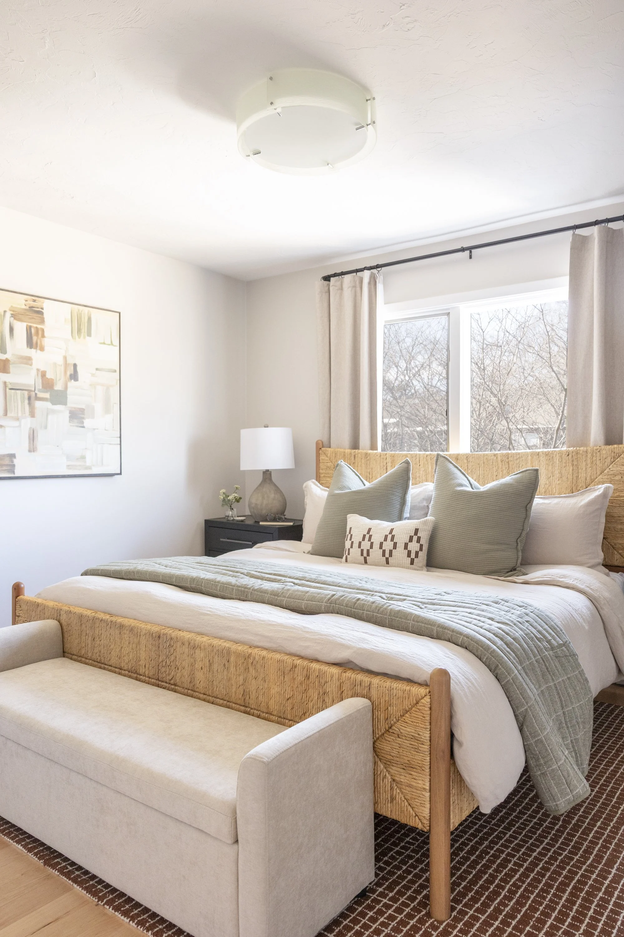 The bedroom features a bed with multiple pillows, a beige upholstered bench at the foot of the bed, a nightstand with a large lamp, a framed abstract painting on the wall, a window with beige curtains, and a textured ceiling light fixture. There is a