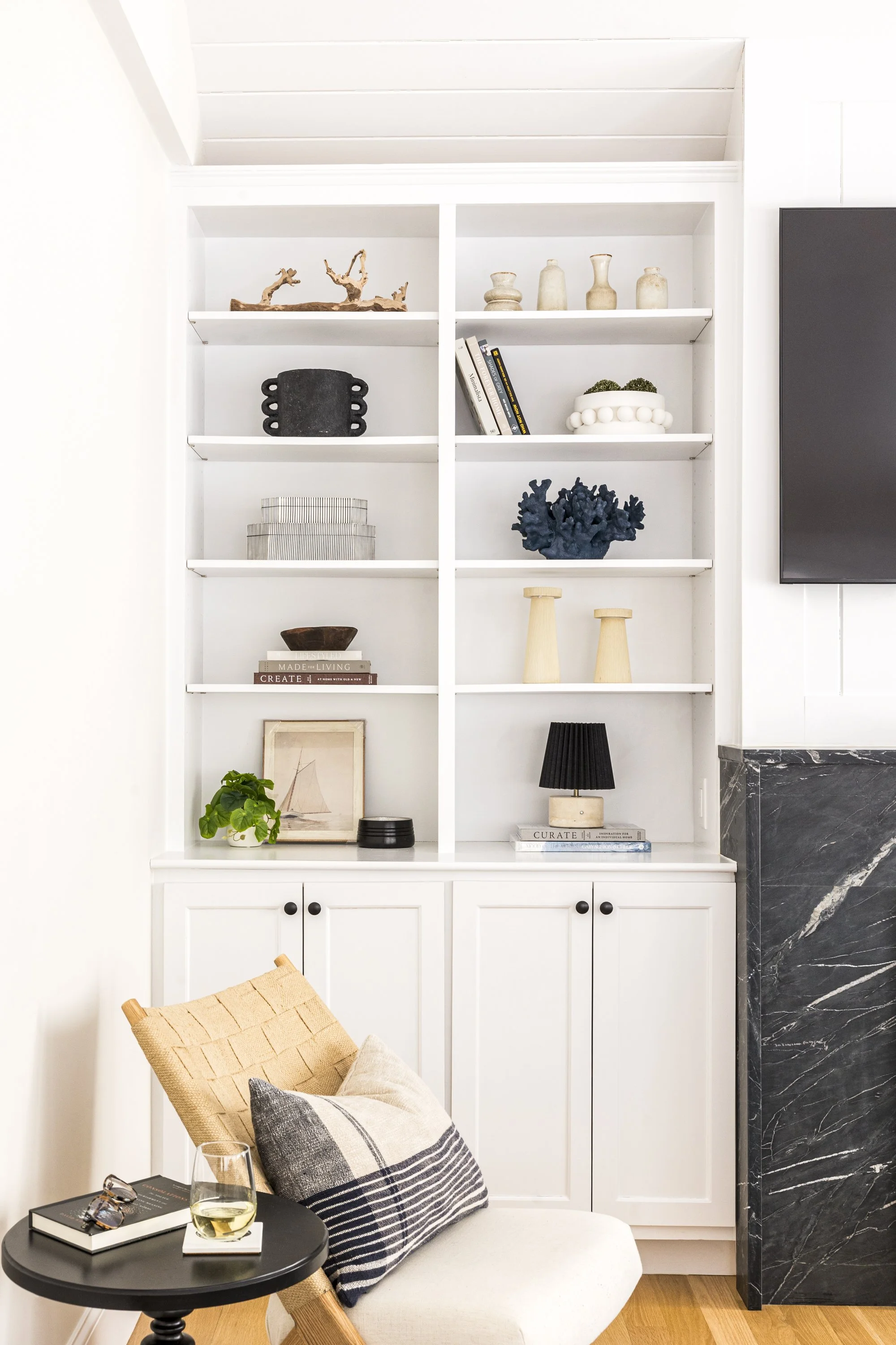Living room with white built-in shelves holding decorative items, a beige chair with a black and white pillow, a small black side table with wine, glasses, a book, and sunglasses, and a dark marble fireplace surround.
