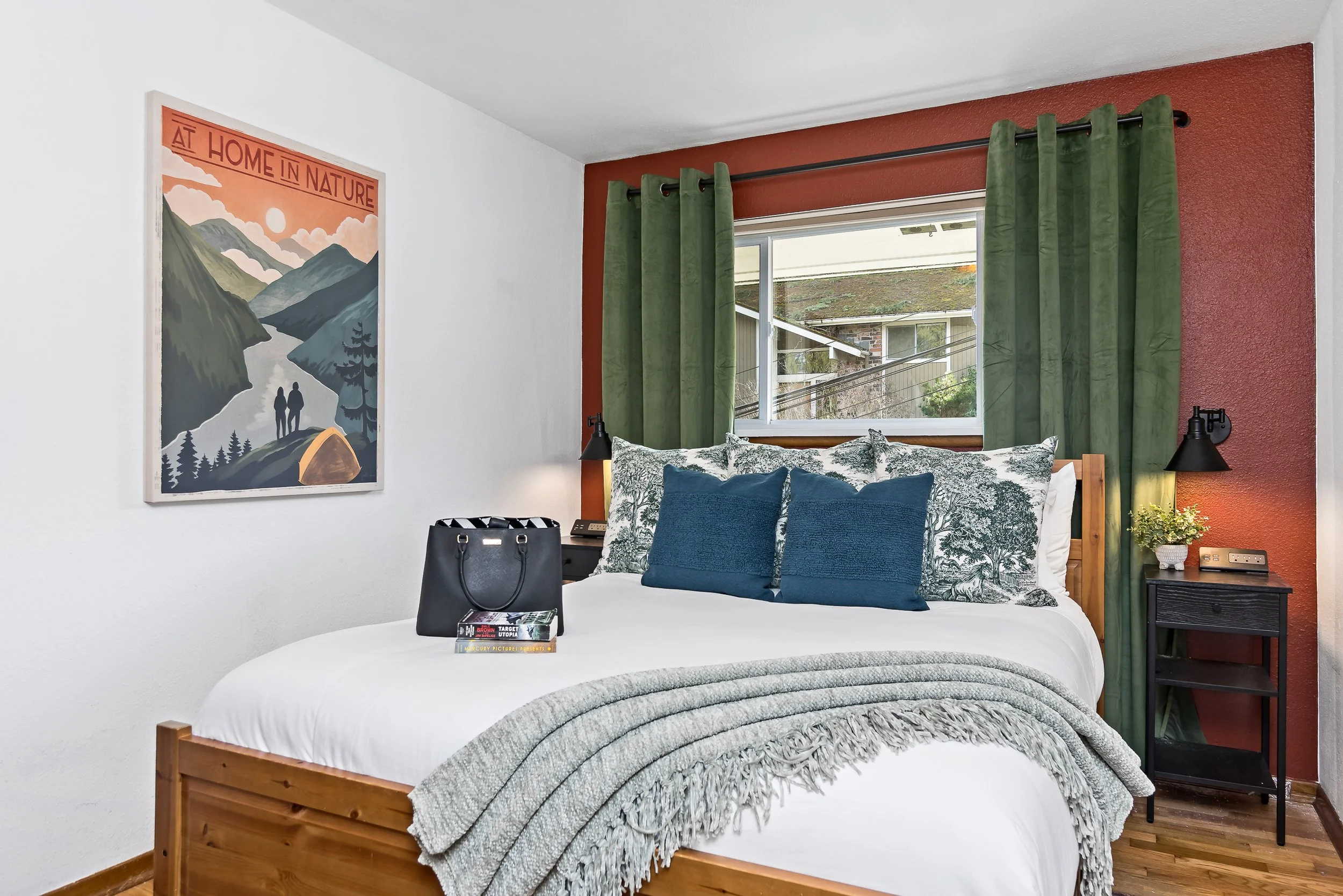 A bedroom with a wooden bed frame, white bedding, blue and patterned pillows, a bedside table with a black lamp, and green curtains in front of a window showing a house and trees outside. A framed poster of mountains, a sunset, and the phrase 'At Hom