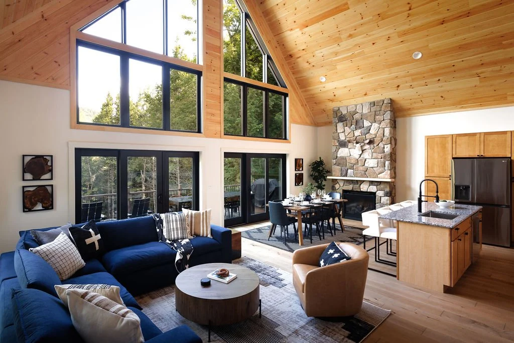 Interior of a modern living room with large windows, wooden ceiling, stone fireplace, and a cozy seating area including a blue sofa, beige armchair, and dining table.