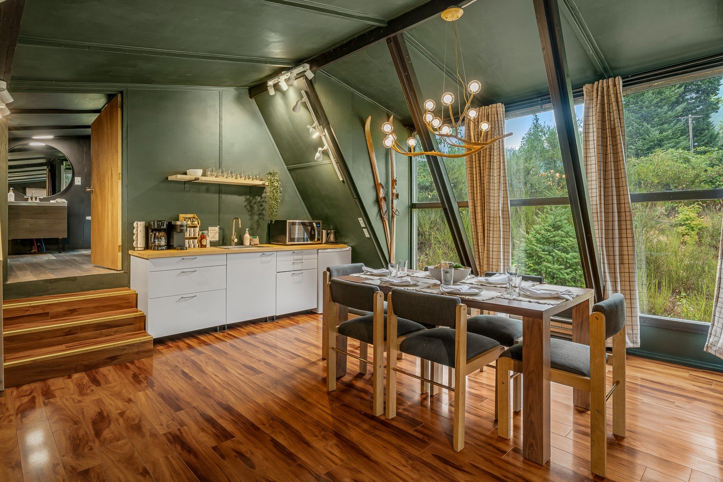 A dining area in a modern cabin with a wooden table set for a meal, six chairs with gray cushions, large windows with curtains showing green trees outside, a chandelier hanging from the ceiling, a kitchenette with white cabinets, a coffee maker, micr