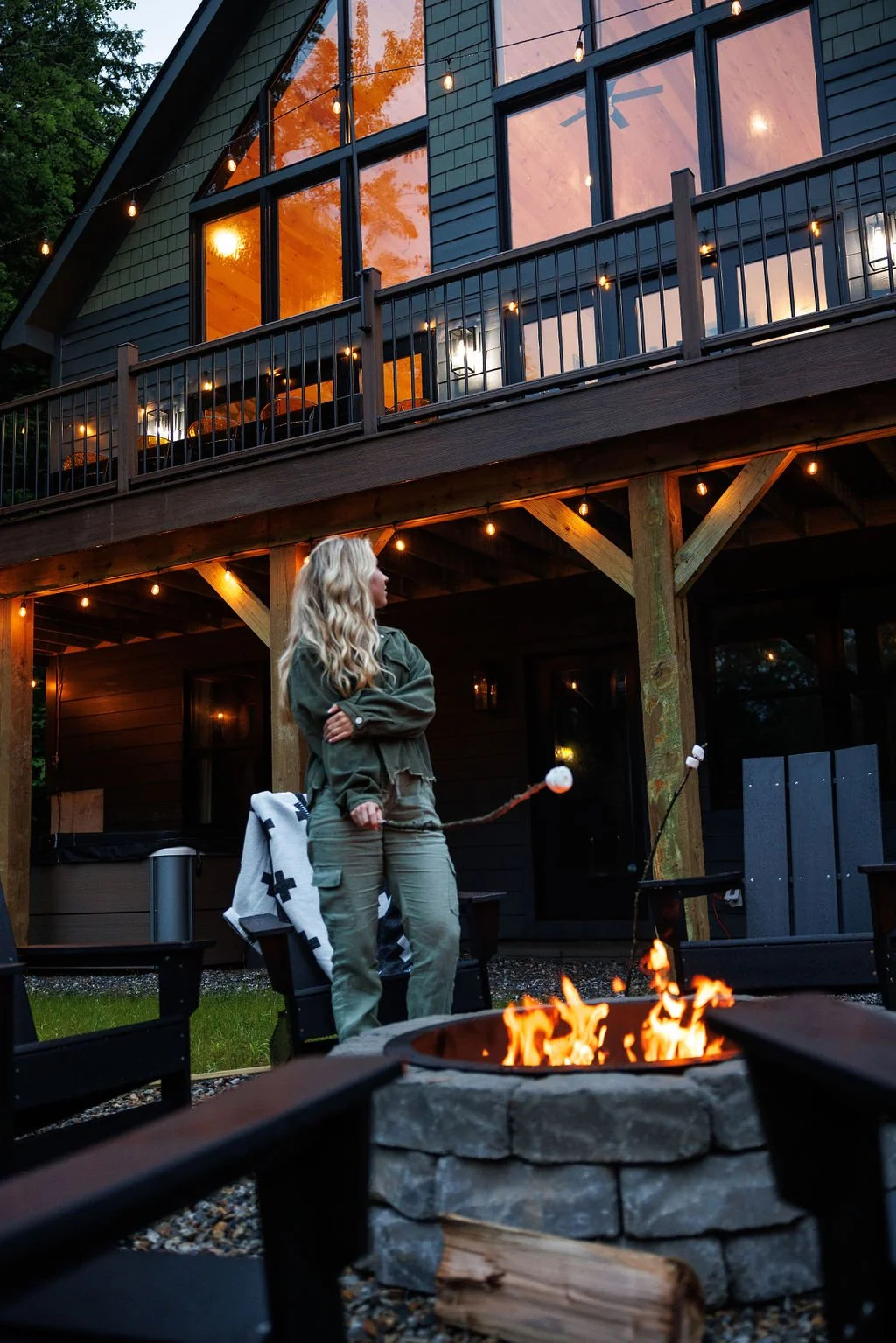 A woman with long blonde hair wearing a green jacket and cargo pants stands near a fire pit in a backyard at dusk. She is holding a marshmallow on a stick. There is a two-story house with large windows and a balcony decorated with string lights in th