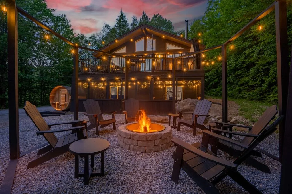 Outdoor backyard seating area with a fire pit, Adirondack chairs, string lights, and a house with a deck surrounded by trees at dusk.