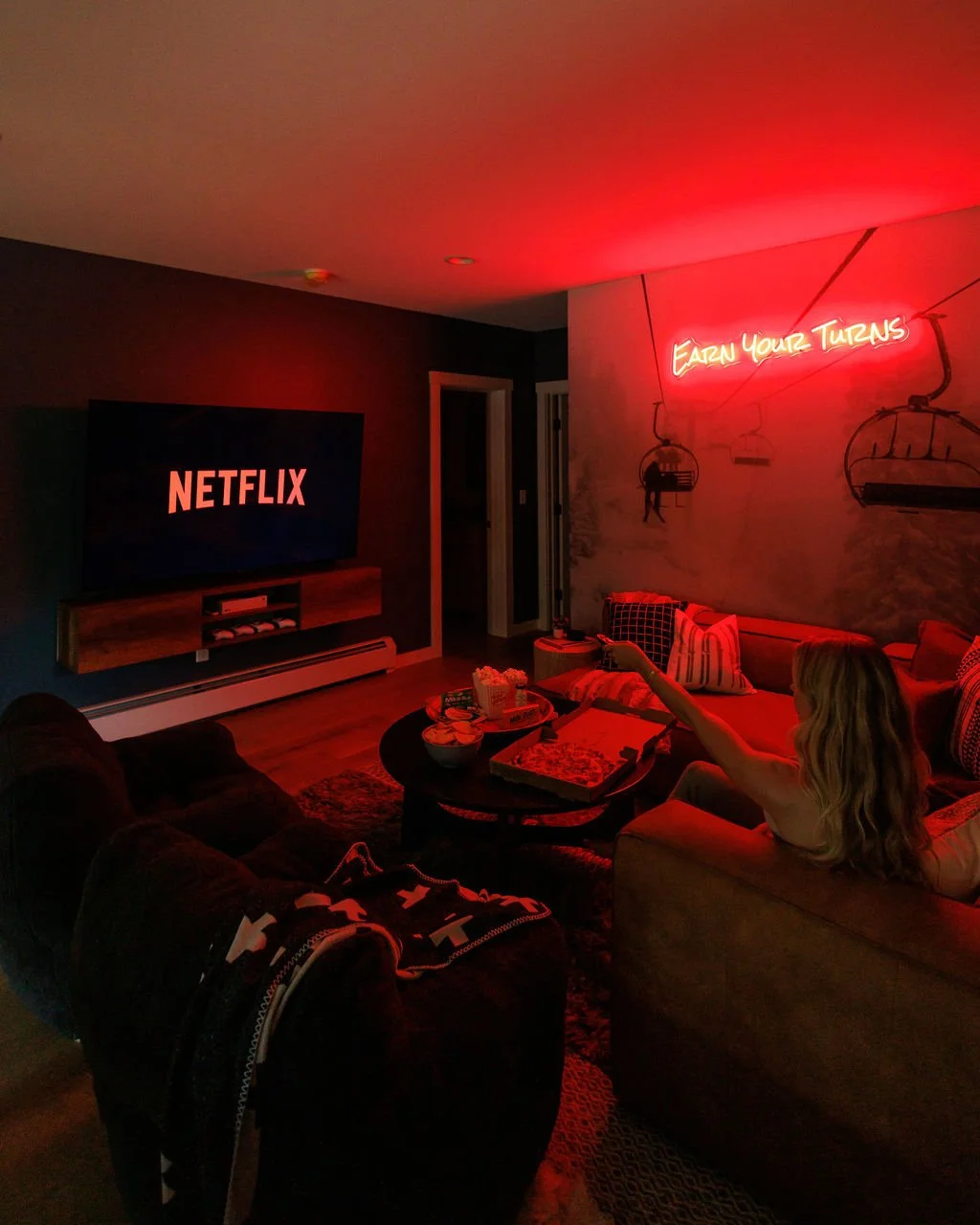 A cozy living room illuminated by red neon lighting with a neon sign on the wall that says 'Earn Your Turns.' A woman is sitting on a couch, reaching for snacks on a coffee table. A large television on the wall displays the Netflix logo.