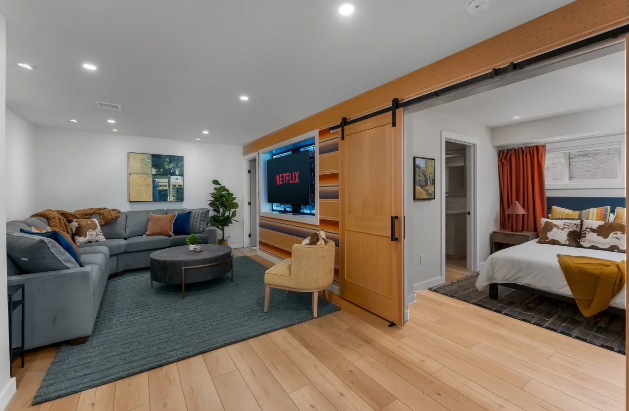 A modern living room with a light gray sectional sofa, a round coffee table, and a yellow accent chair. A wall-mounted TV screens Netflix, with a sliding barn door on a wooden track separating the living area from a bedroom. The bedroom has a bed wit