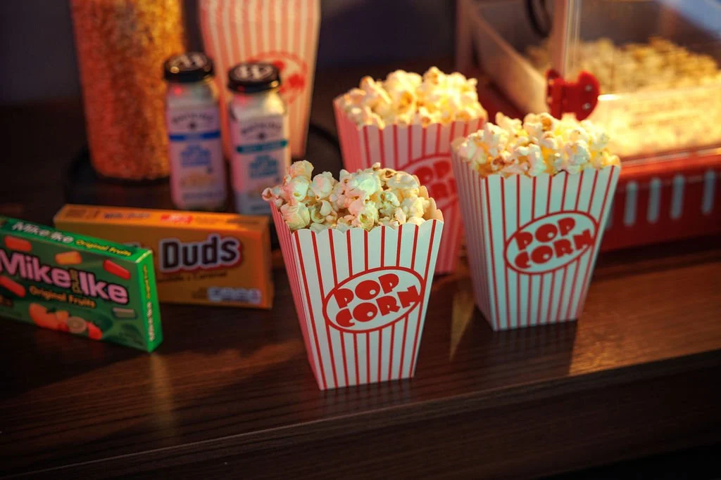 Three containers of popcorn on a dark wooden table, with various snack items and drinks in the background.