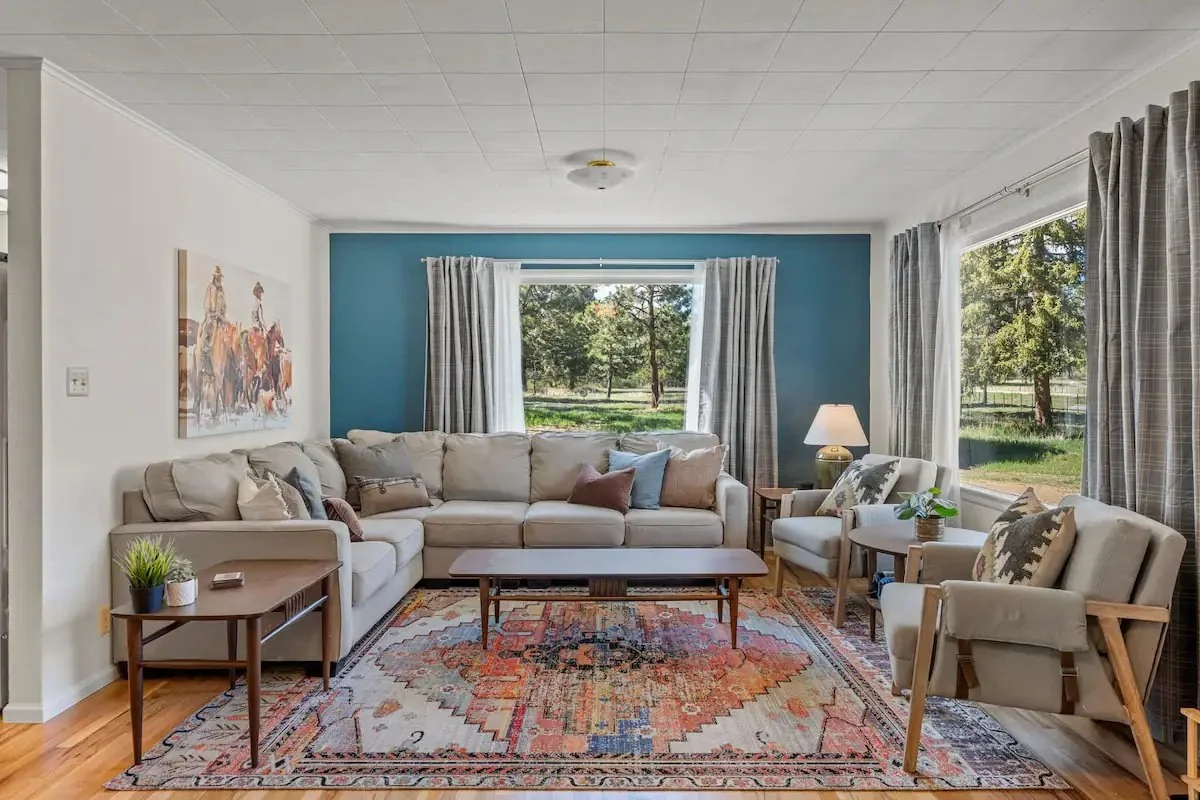 Living room with a large beige sofa, two armchairs, a wooden coffee table, side tables with plants, a colorful area rug, and large windows with curtains showing a green outdoor landscape.