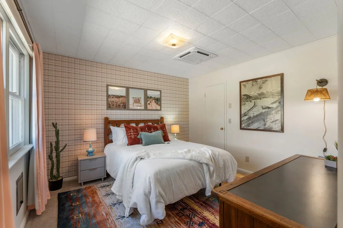 A bedroom with a bed, nightstand, and artwork, featuring a cactus plant next to a window with pink curtains.