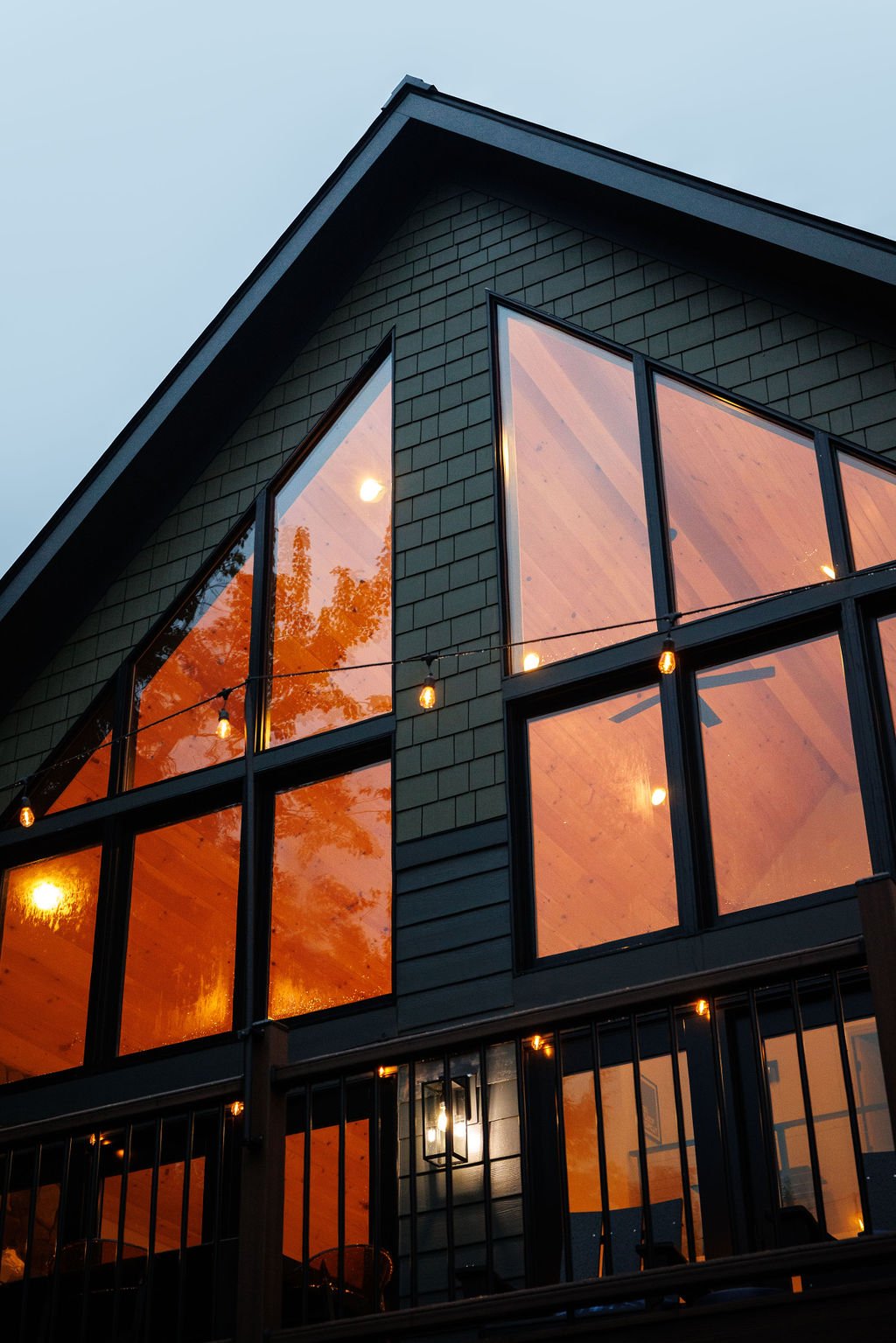 A modern house with large triangular and rectangular windows reflecting orange evening sky and trees, string lights hanging outside.