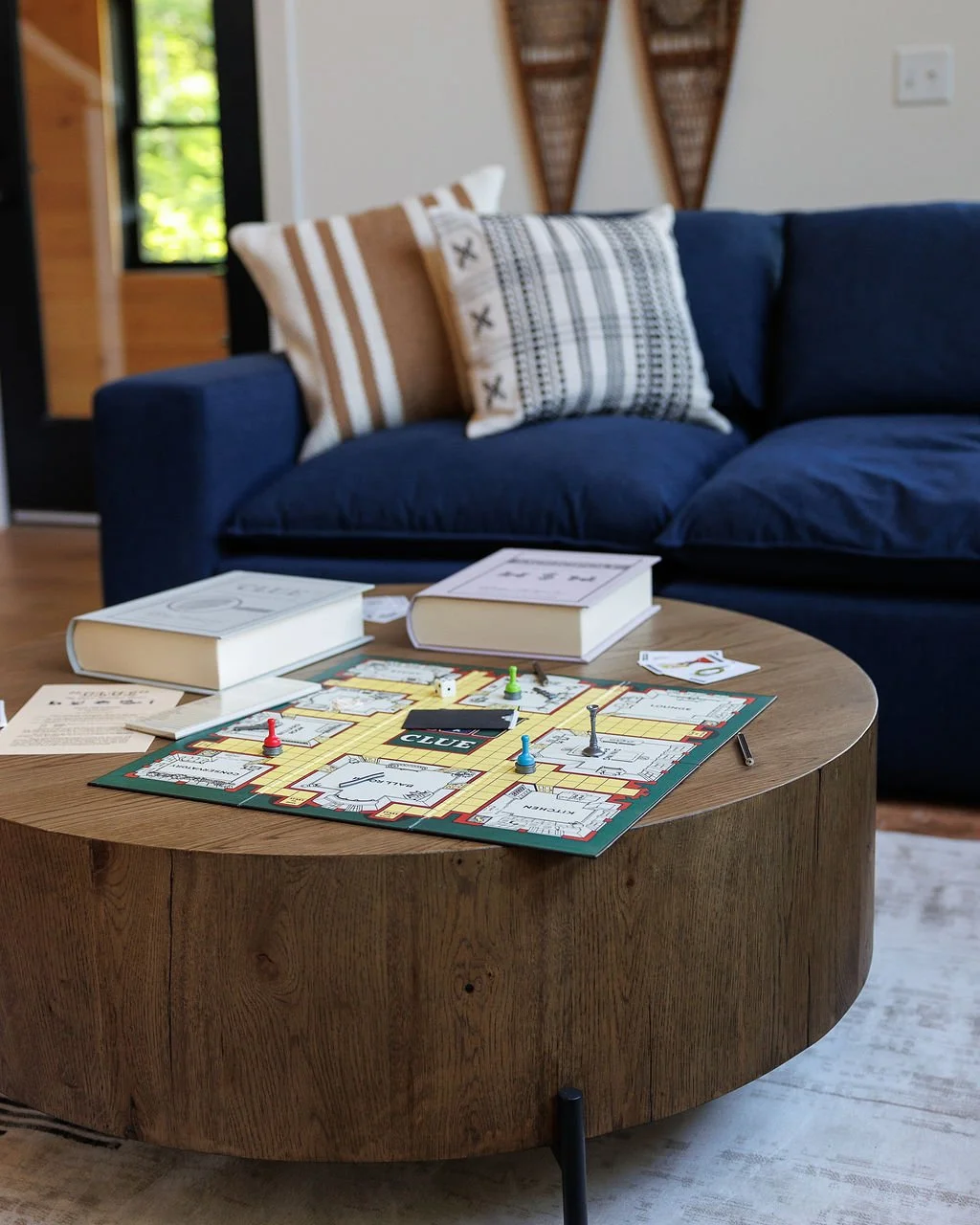 Living room with a blue sofa, a wooden coffee table with a board game, books, and game pieces, and patterned pillows, with wall art and a window in the background.