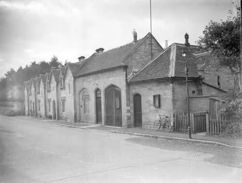 Black and white photo of a row of old buildings on a street, with a bicycle parked near a fence and trees in the background.