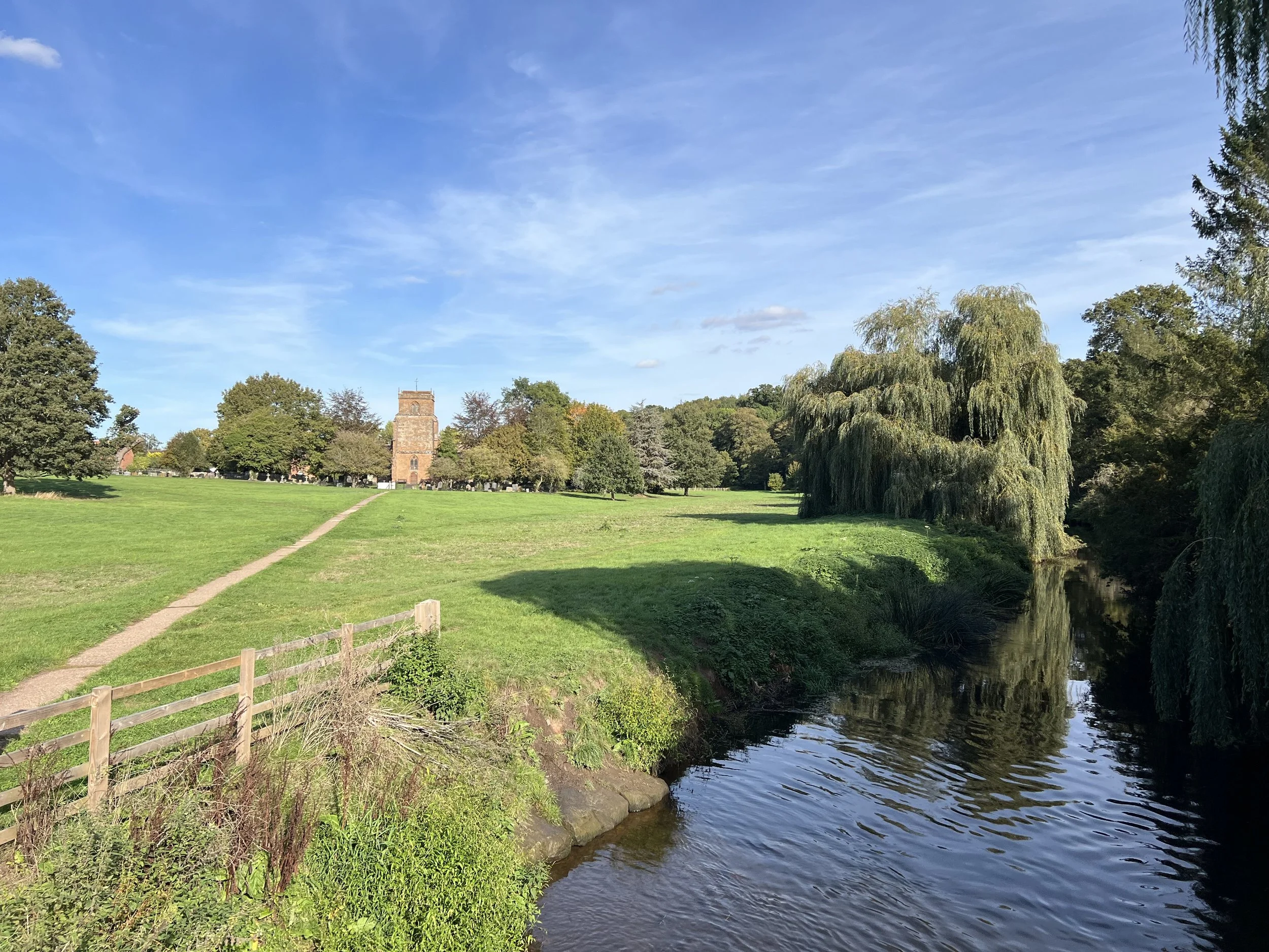 A park scene with a green grassy area, a walking path, and a small river with gentle ripples. There are trees and a brick building or church in the background under a blue sky with some clouds.