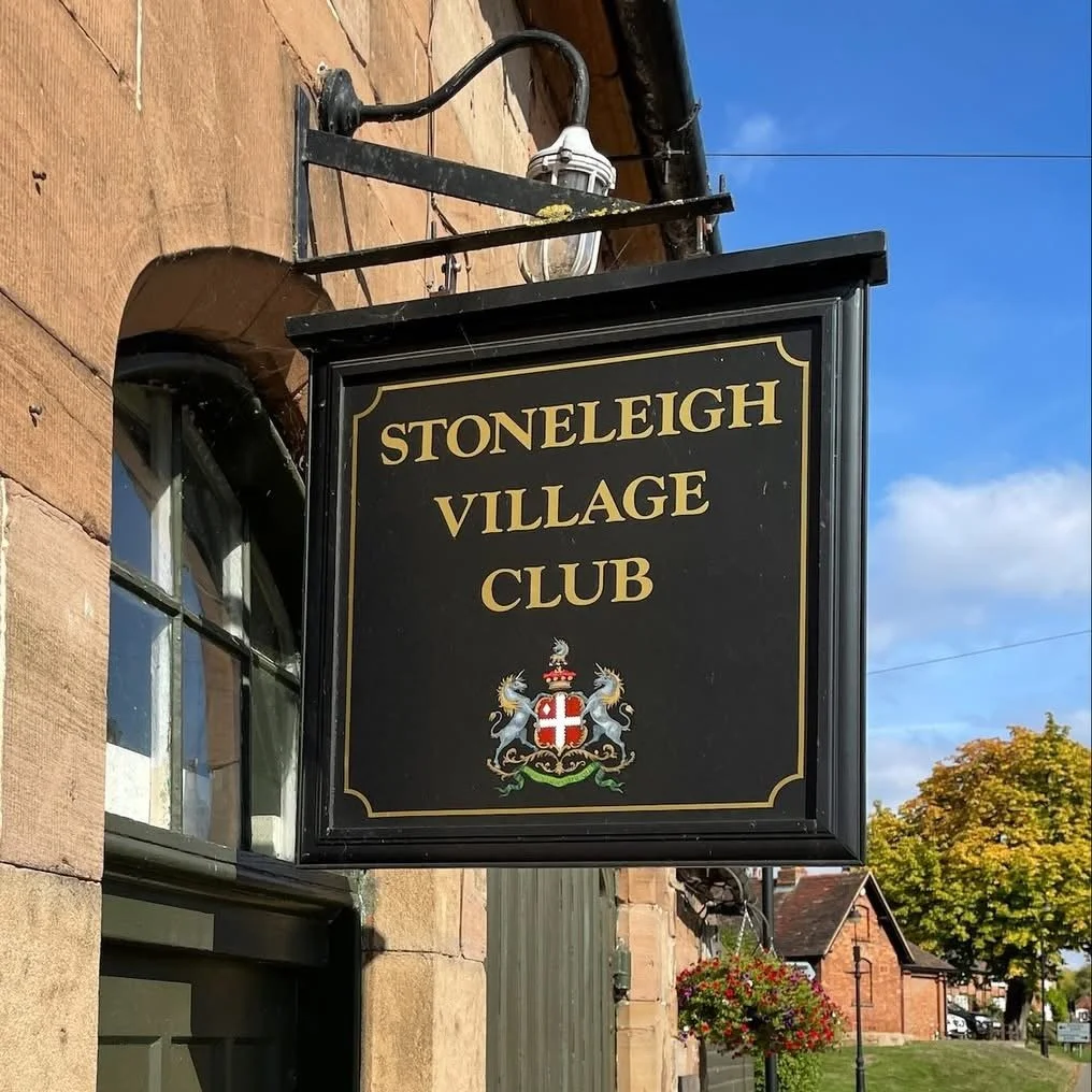 Black sign with gold lettering that reads 'Stoneleigh Village Club' with a crest below, hanging outside a stone building with a window, in a village street with trees and houses.