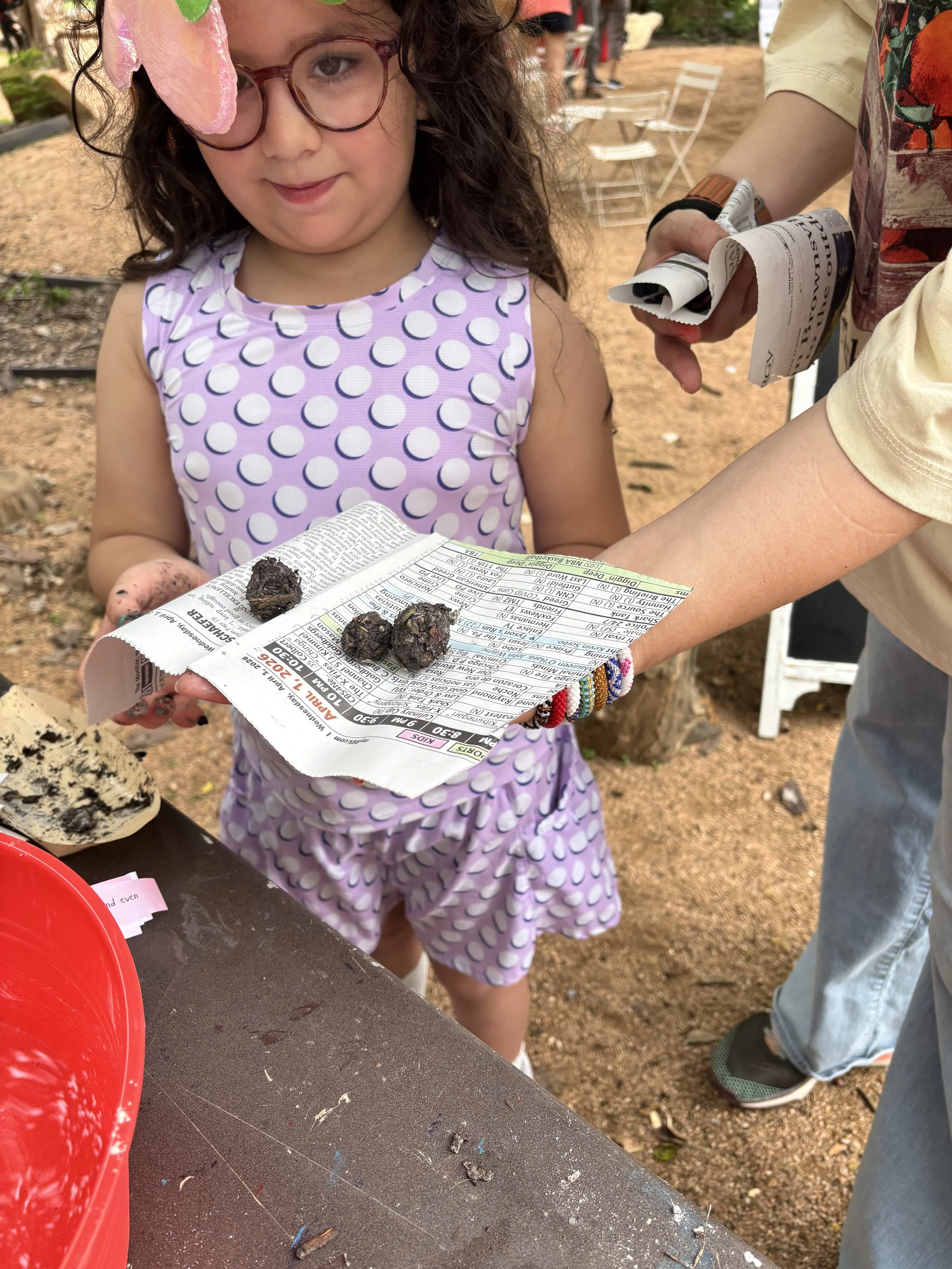 A young girl with glasses and curly hair wearing a purple polka dot dress holding a rolled newspaper with three seed pods on top, standing at an outdoor event or fair.