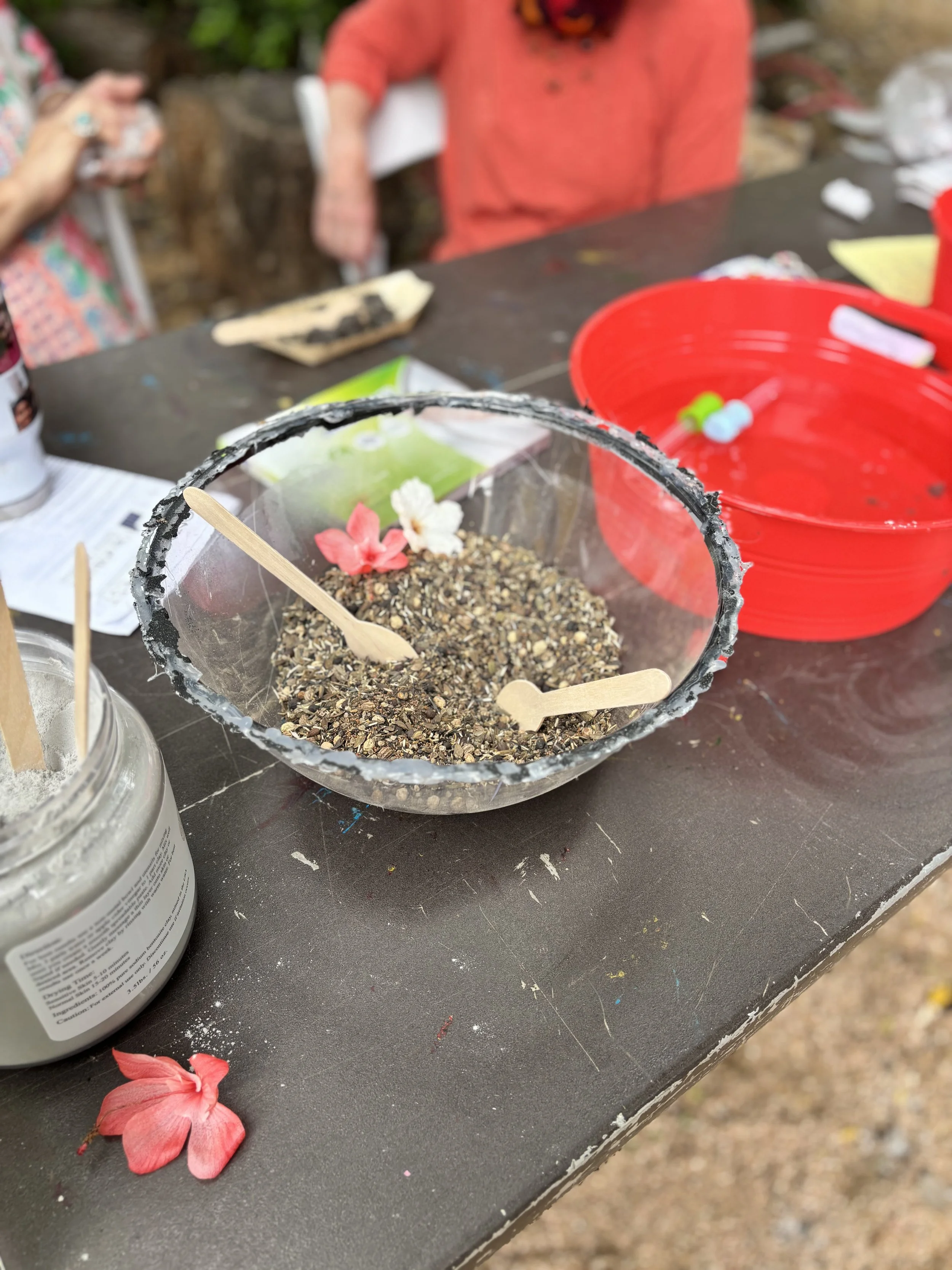 Close-up of a black container filled with dirt, small pebbles, and decorative pink and white flowers, with a wooden stick inside. Items are on a black table, including a red bowl, a jar of white powder, and other objects, with blurred people in the b