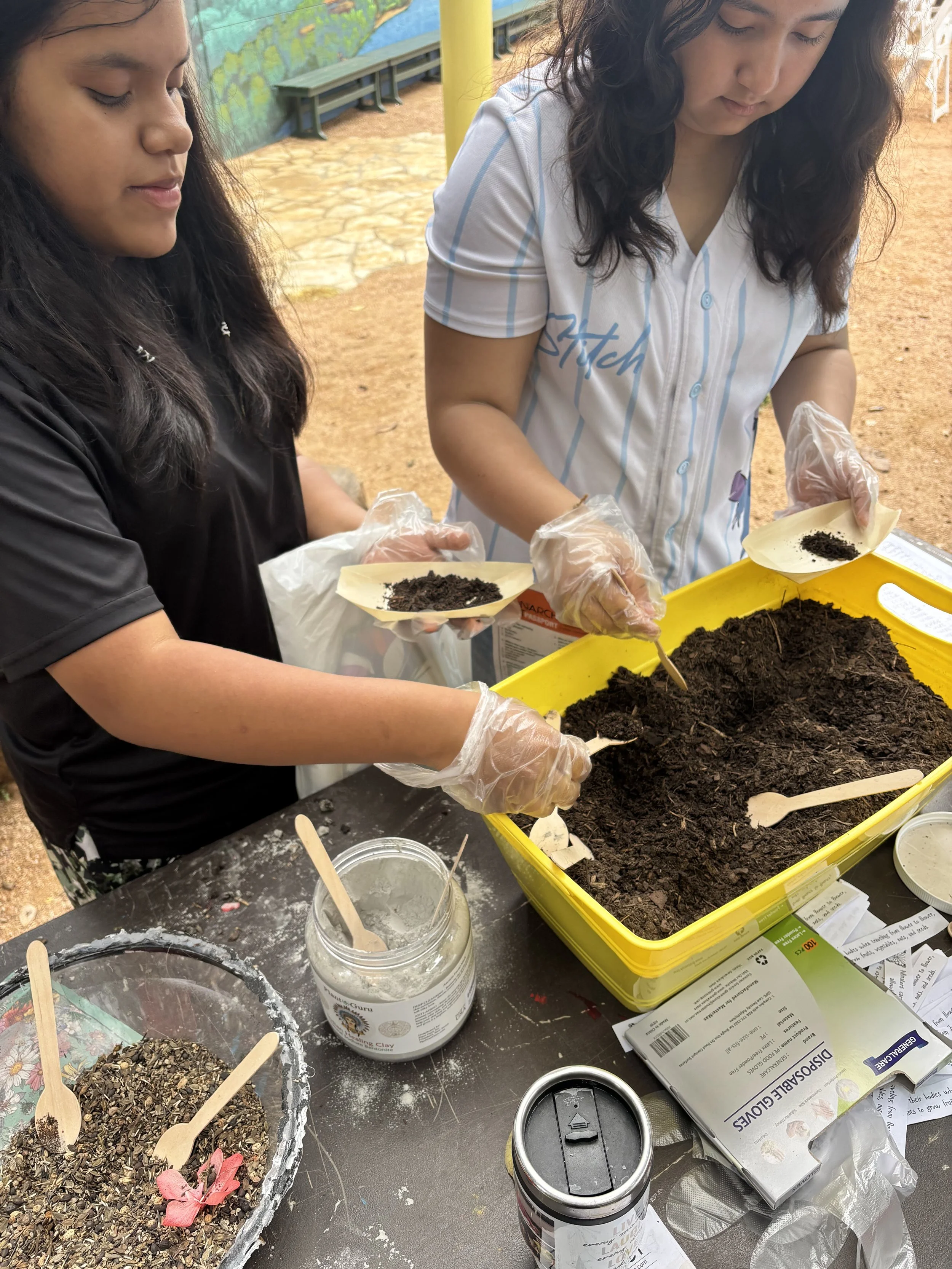 Two girls planting seeds in soil at a table with gardening supplies, including a bag of potting mix, seed packets, and gardening tools, outdoors.