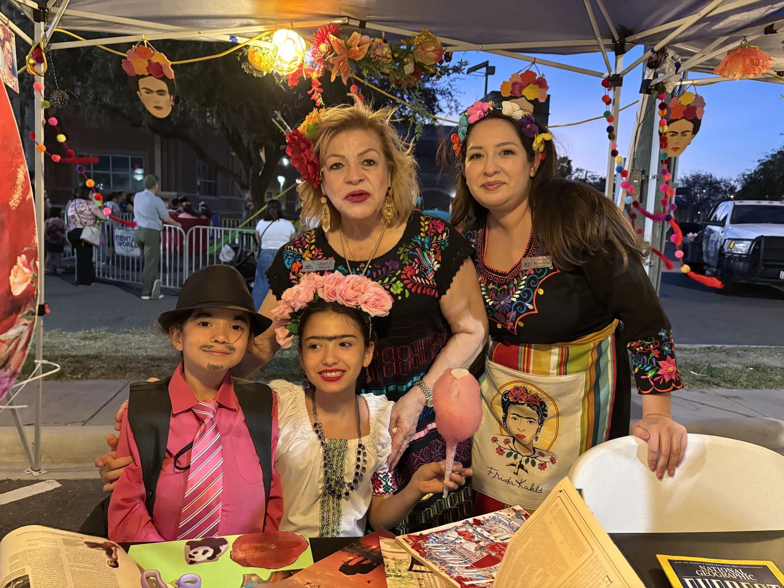 Two women and two children pose for a photo at a festive outdoor event. The women wear colorful traditional outfits and floral headpieces. Colorful decorations and a crowd are visible in the background.