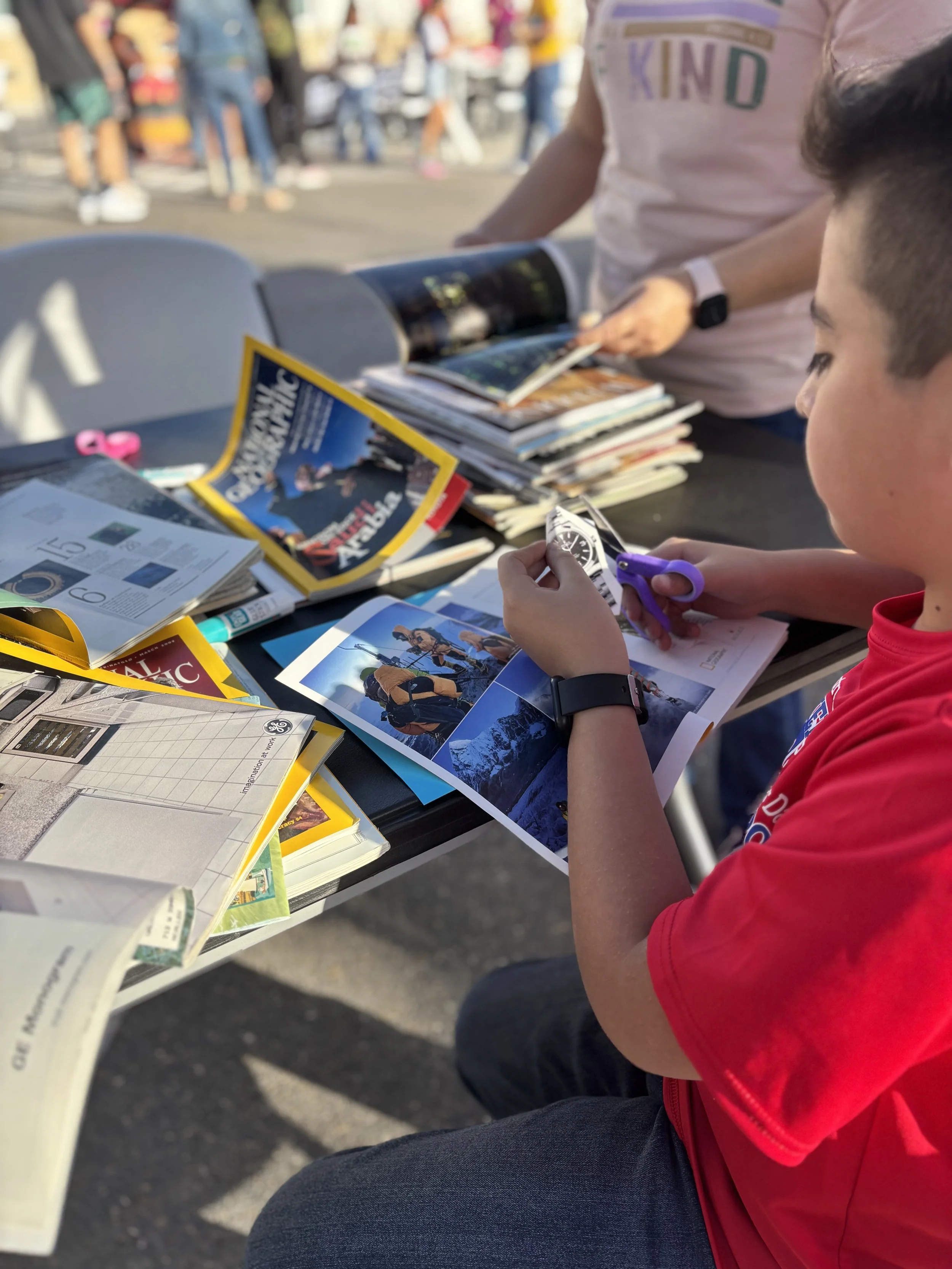 A boy wearing a red shirt and black watch sitting at a table outdoors, looking at a brochure, with magazines and pamphlets spread out on the table, and a person standing next to him flipping through a magazine in the background, with a crowd of peopl