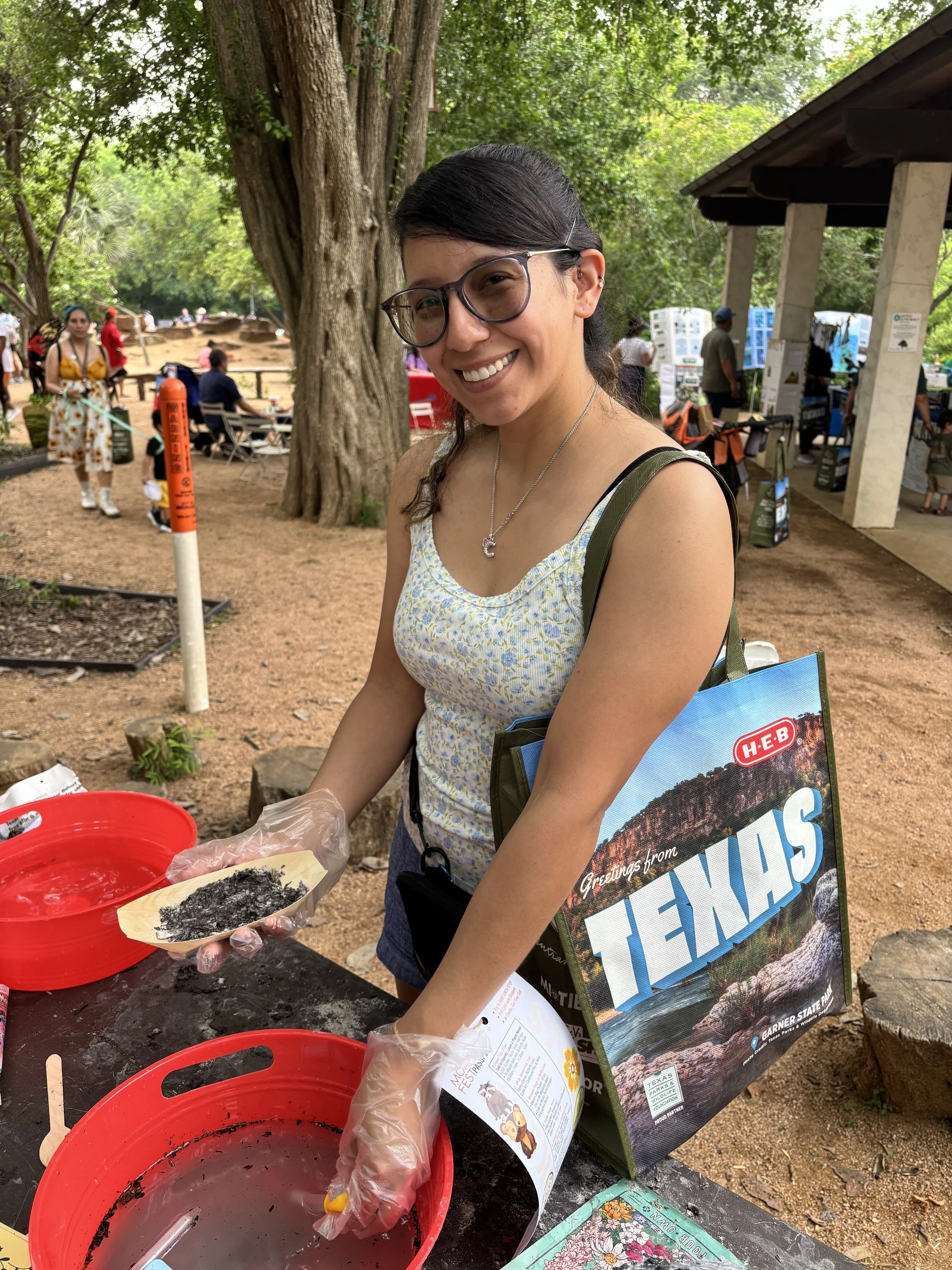 A woman wearing glasses and a yellow floral dress smiling while handling a bamboo plate filled with soil and seeds, outdoors near trees and other people at a community event.