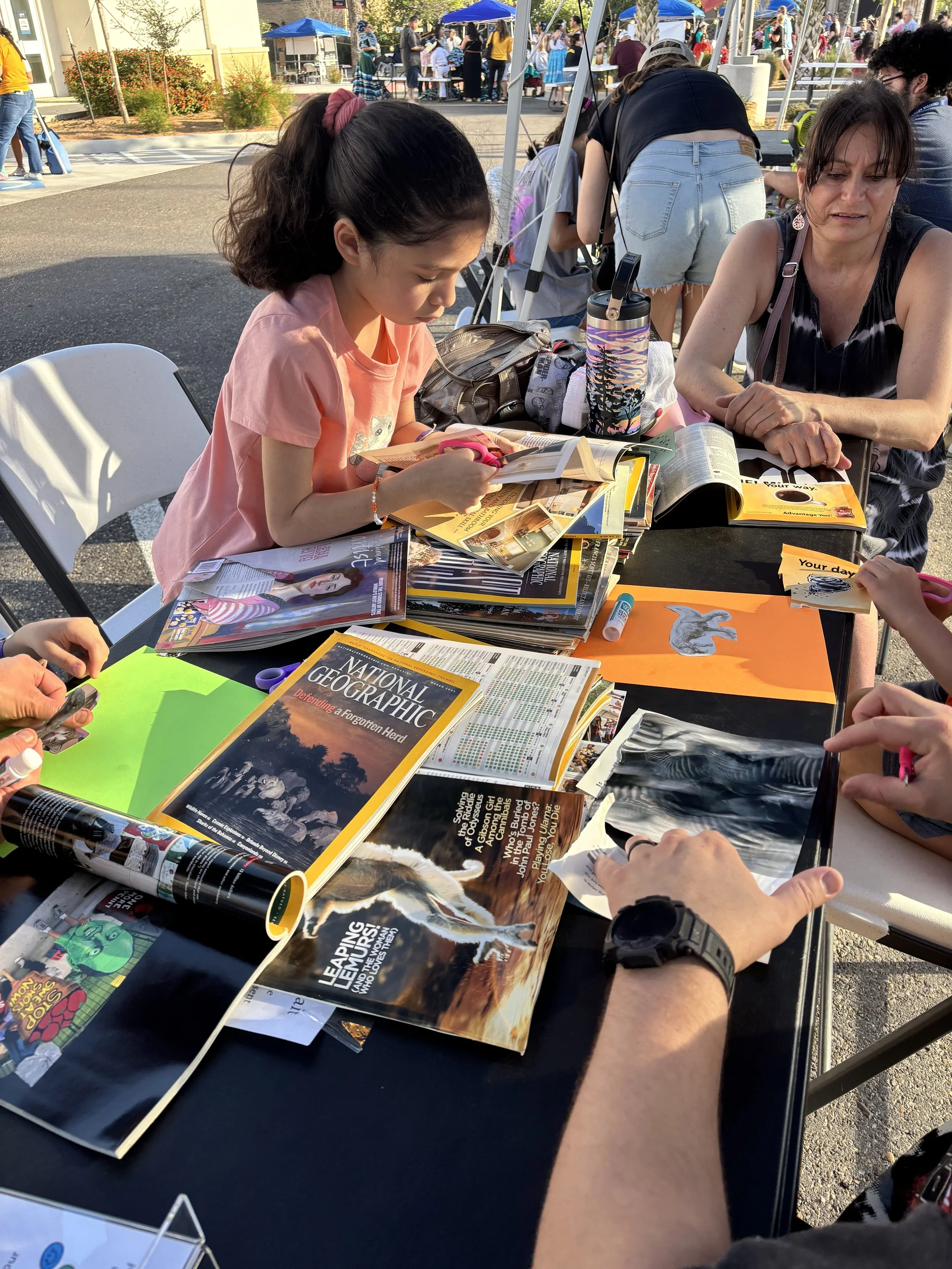 A young girl and an older woman are sitting at a table outdoors, looking through magazines and catalogs. The table is filled with various magazines, including National Geographic. There are other people in the background at an outdoor event, with ten