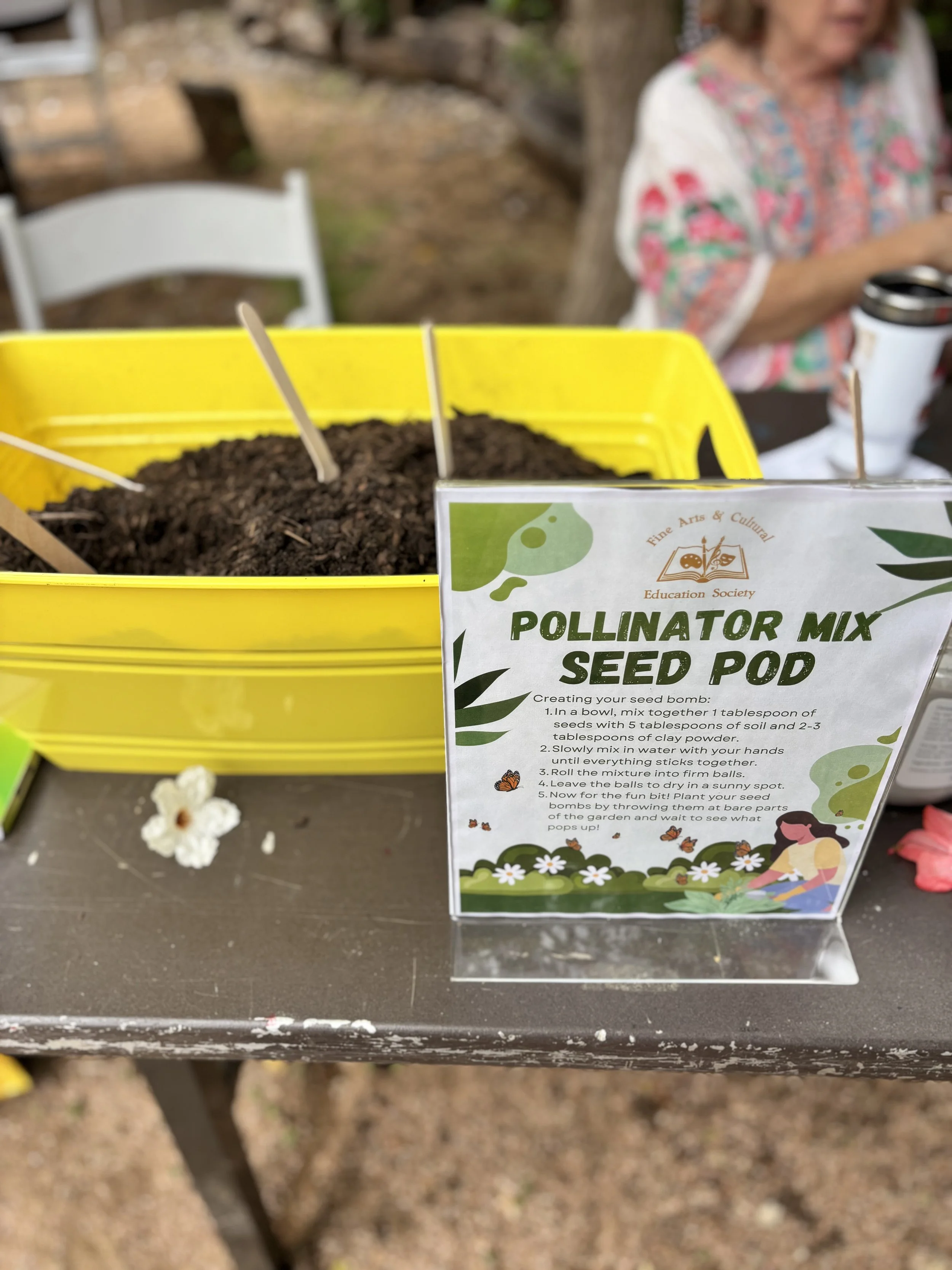 A yellow plastic container filled with soil, labeled 'Pollinator Mix Seed Pod,' on a table outdoors. There is a woman in the background, sitting at a table with a coffee mug, partially out of focus.