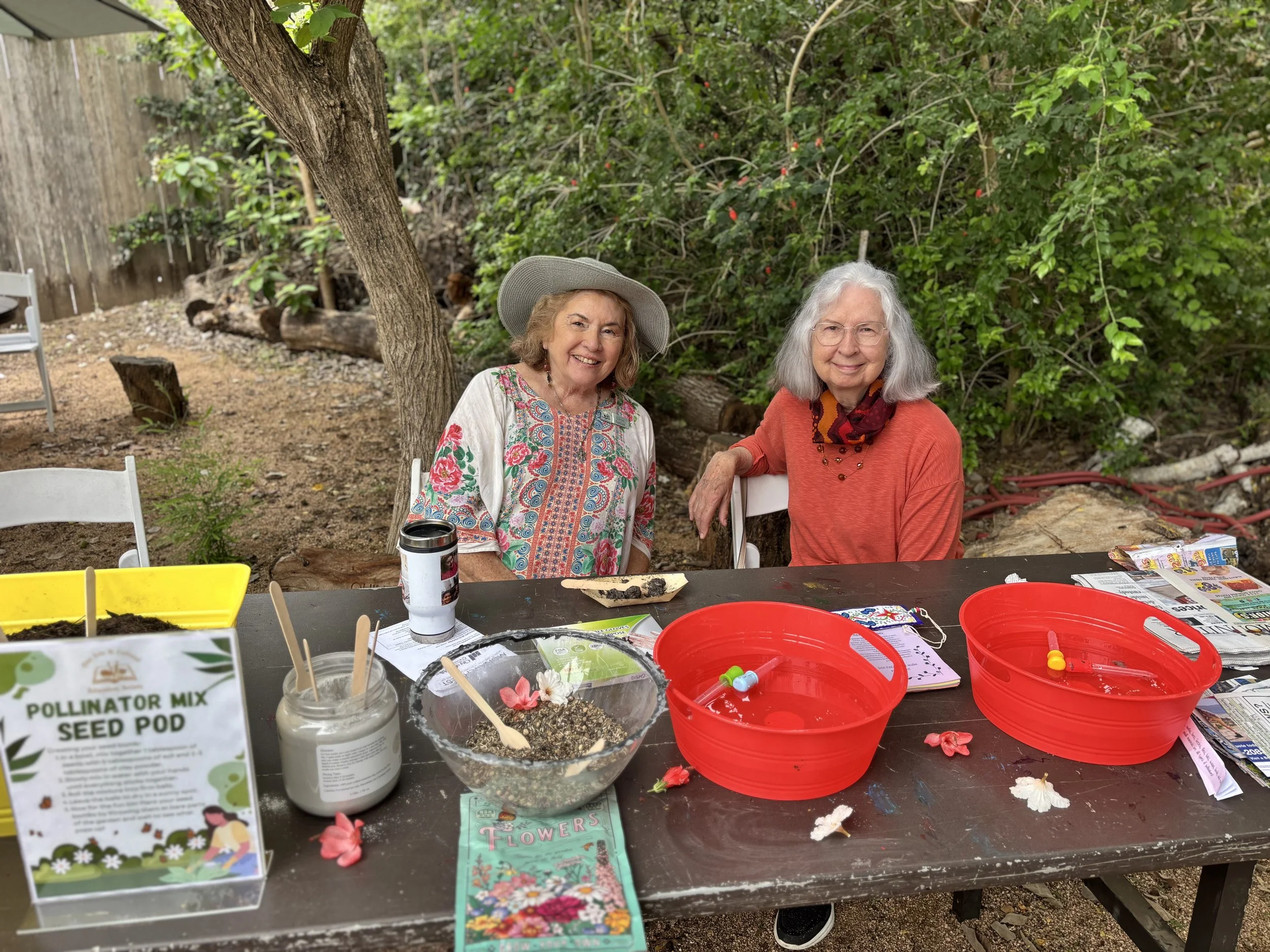 Two elderly women sitting at a table outdoors in a garden, surrounded by trees and greenery, with gardening supplies and seed packets on the table.