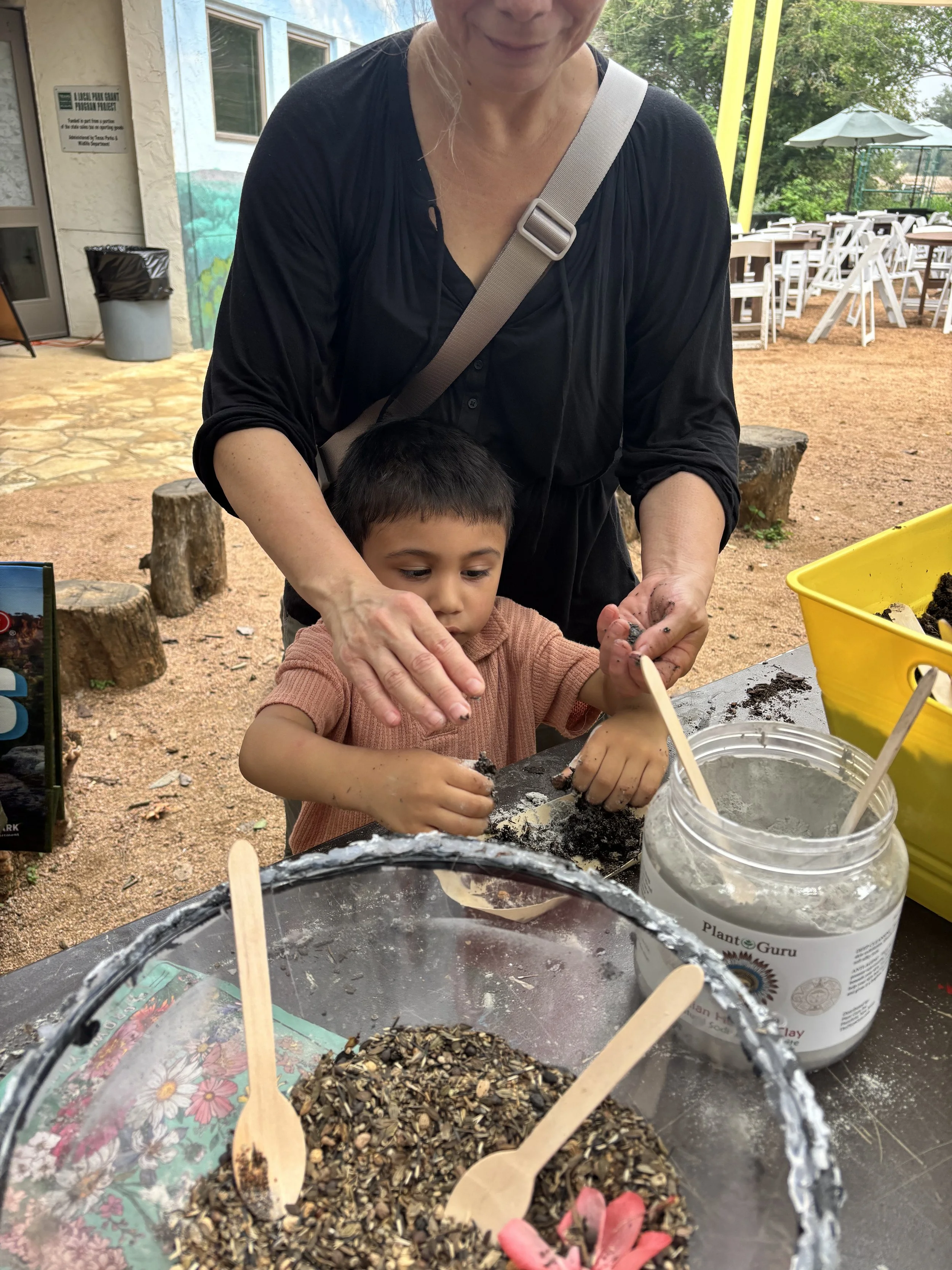 A woman and a young boy planting seeds outdoors. The woman is guiding the boy who is handling soil or seeds, with garden supplies and a yellow container nearby, and a background of trees and outdoor furniture.