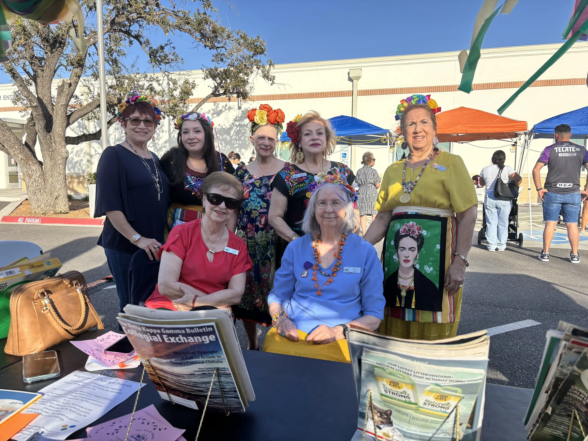 Group of women wearing traditional Mexican attire and flower crowns, standing outside at a cultural event, with tents and other people in the background.