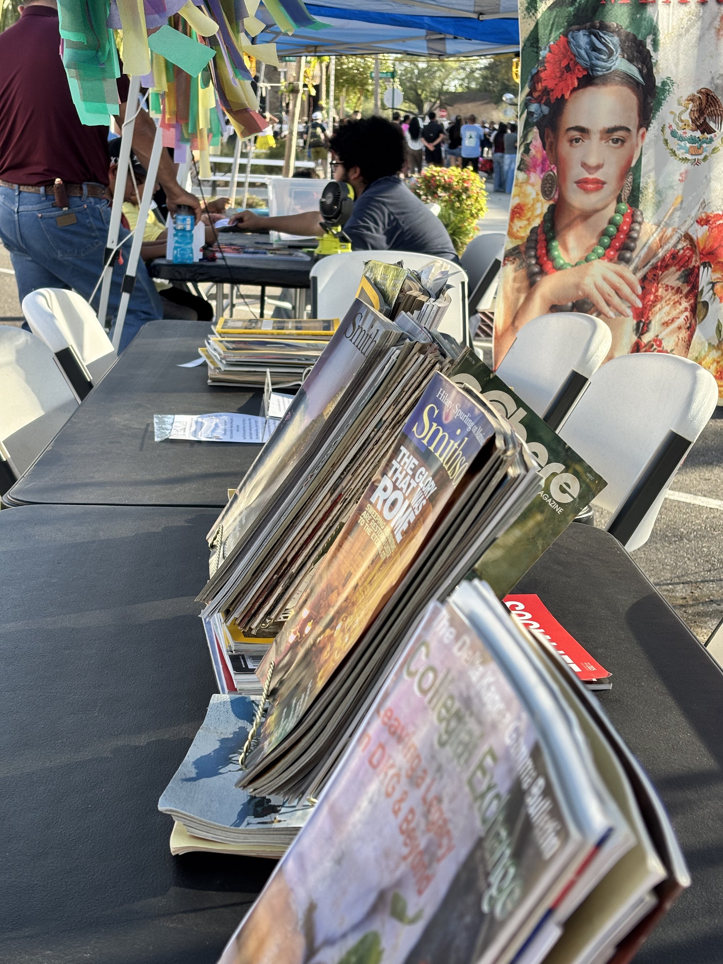 Table of magazines at an outdoor event, with people and booths in the background, and a large poster of Frida Kahlo.