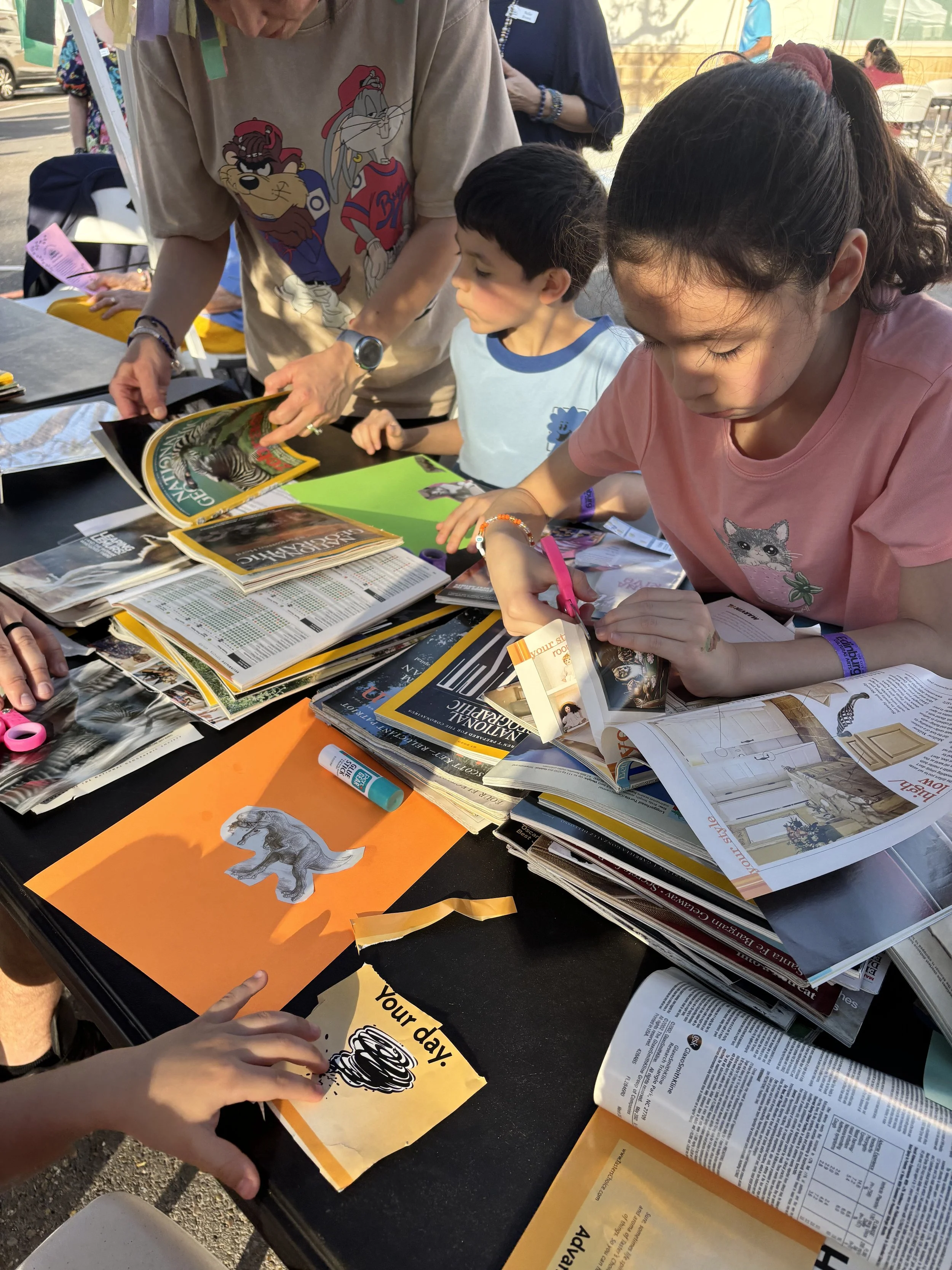 Children and adults gathered around a table with various magazines, maps, and papers, engaged in arts and crafts activities outdoors, with some cutting and coloring papers.