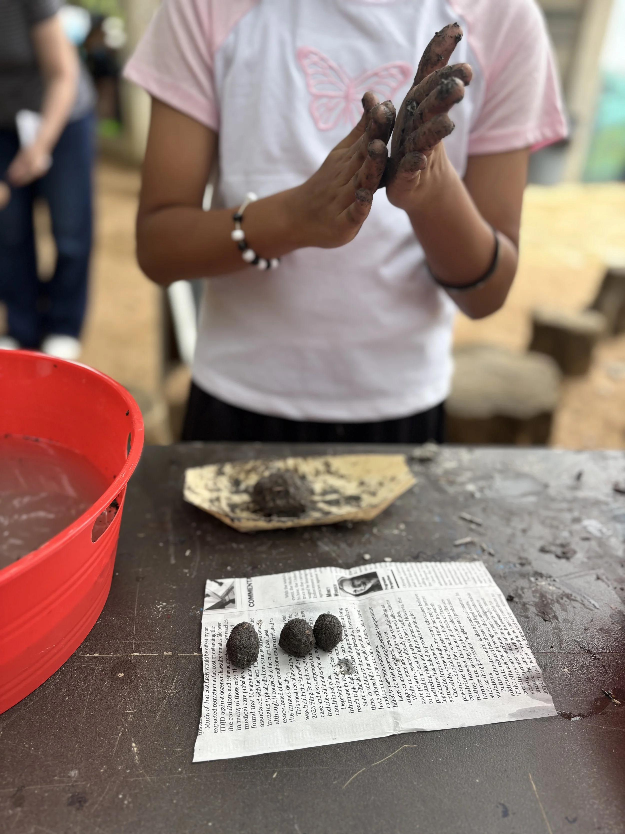 Person with dirt-stained hands working with small pods of soil on a newspaper-covered surface, with a red container nearby.