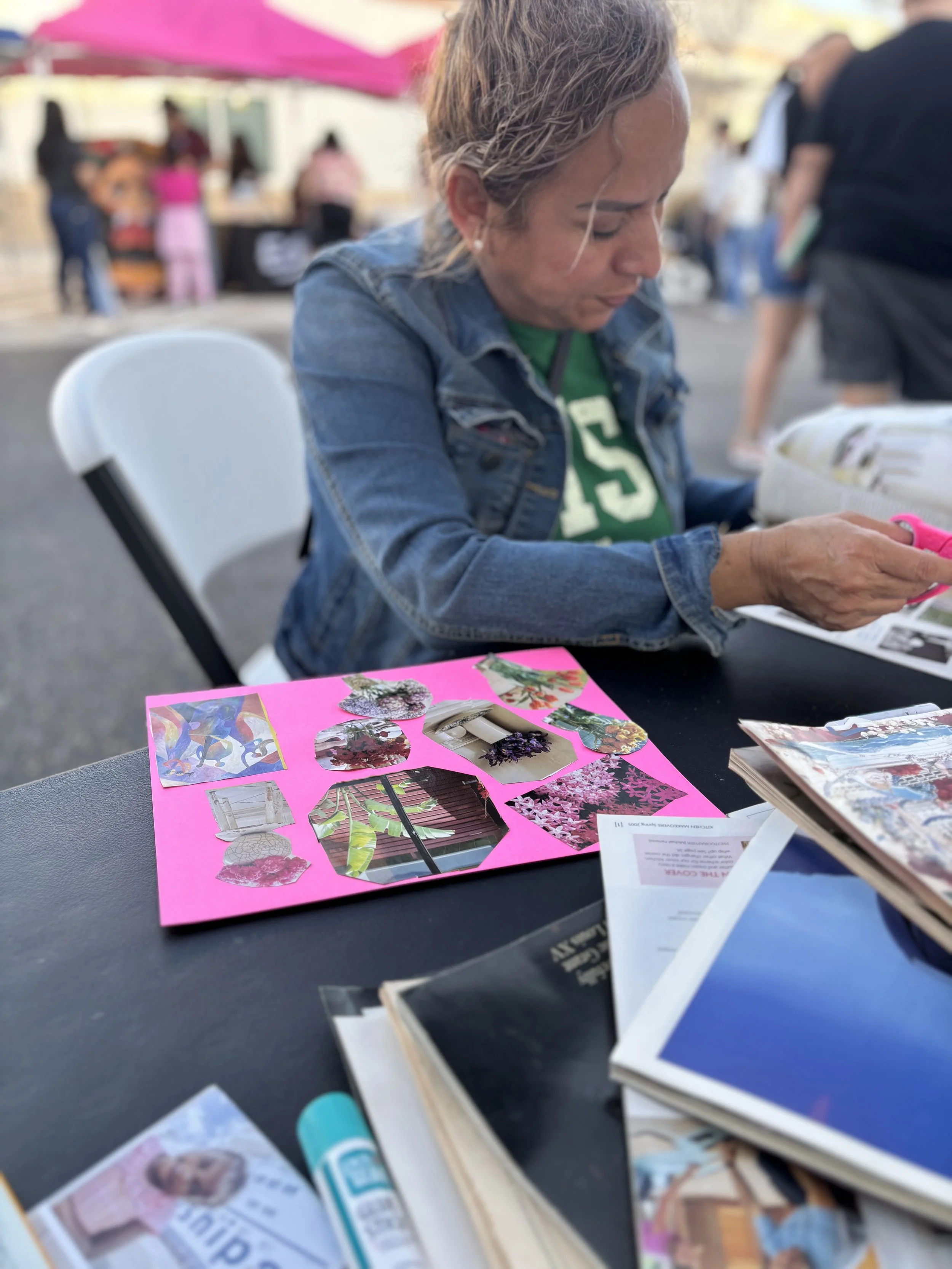 A woman sitting at a table outdoors, looking through magazines. In the foreground, there are more magazines and colorful photo collages on pink construction paper. There are people in the background under a pink umbrella.