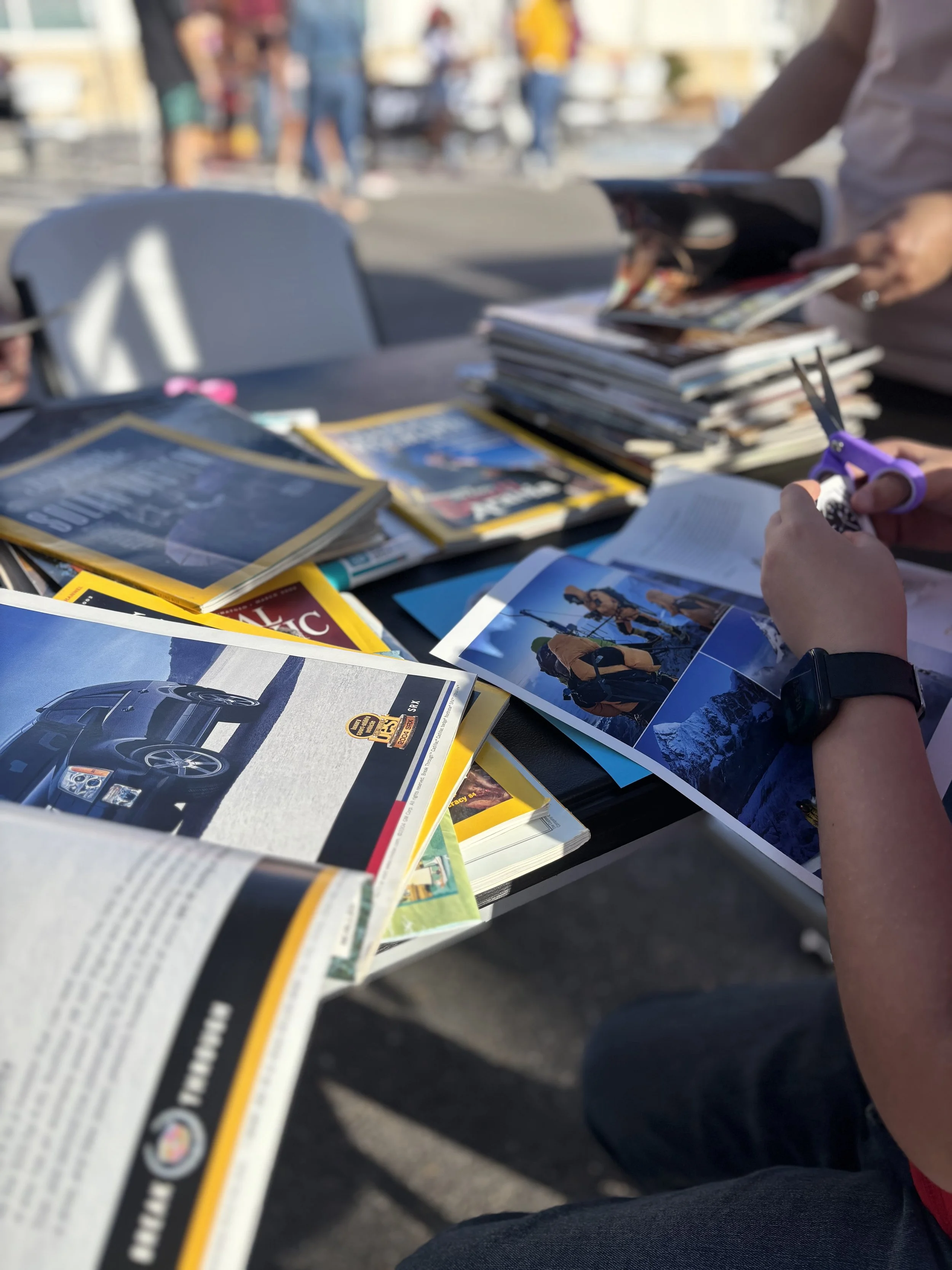 Table covered with magazines, newspapers, and photographs at an outdoor event. People are blurred in the background.